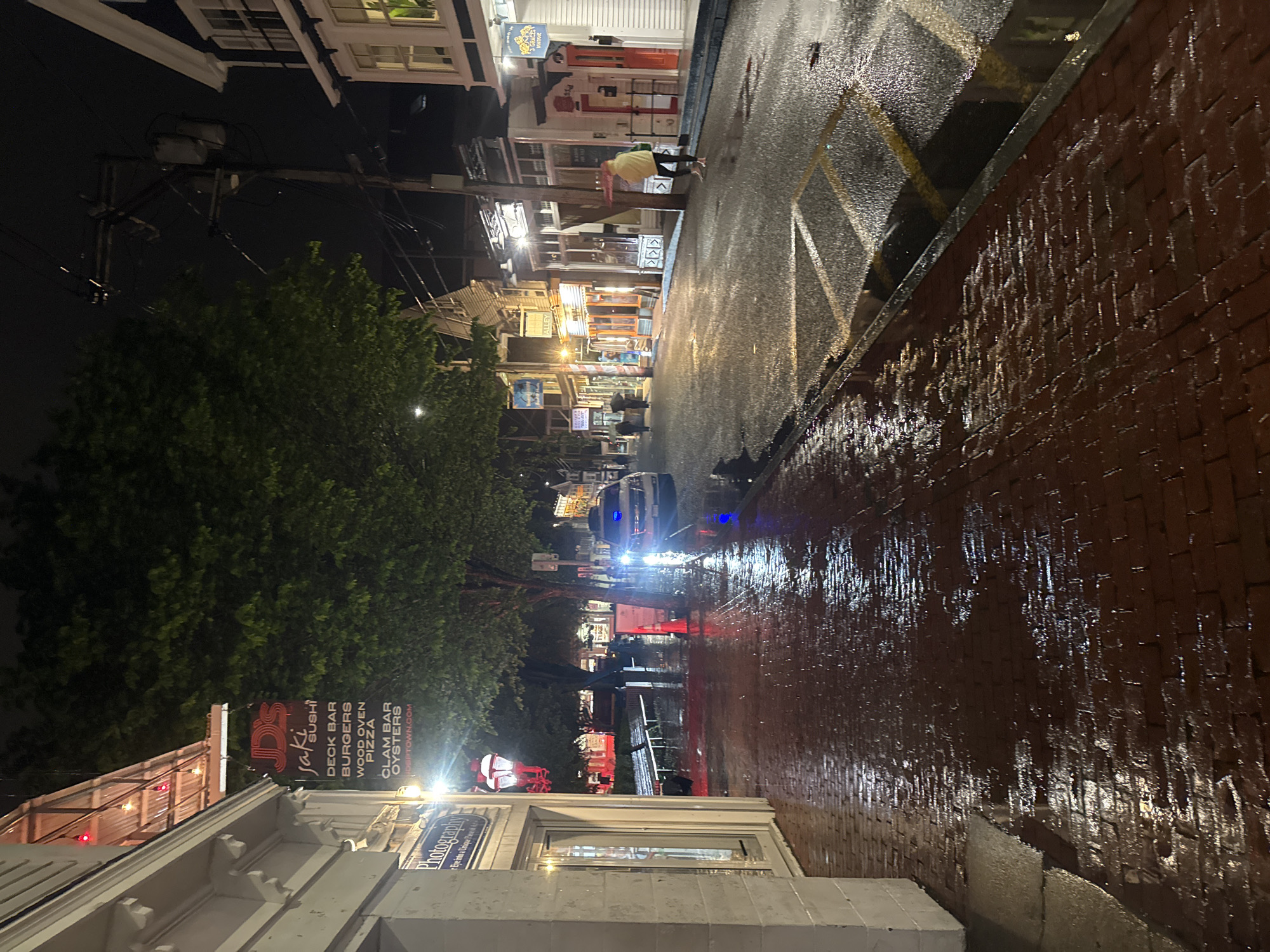 The image depicts a wet street scene at night in Provincetown, Massachusetts. The street appears to be brick-paved and is lined with buildings on both sides. Several people are visible in the distance, some walking with umbrellas. A police car is stationed further down the street with its headlights and blue lights illuminated. Streetlights cast reflections on the wet surface of the street. A restaurant sign, "Jaki's Sushi," is visible on the left side of the image. The overall atmosphere suggests a rainy night in a small town.