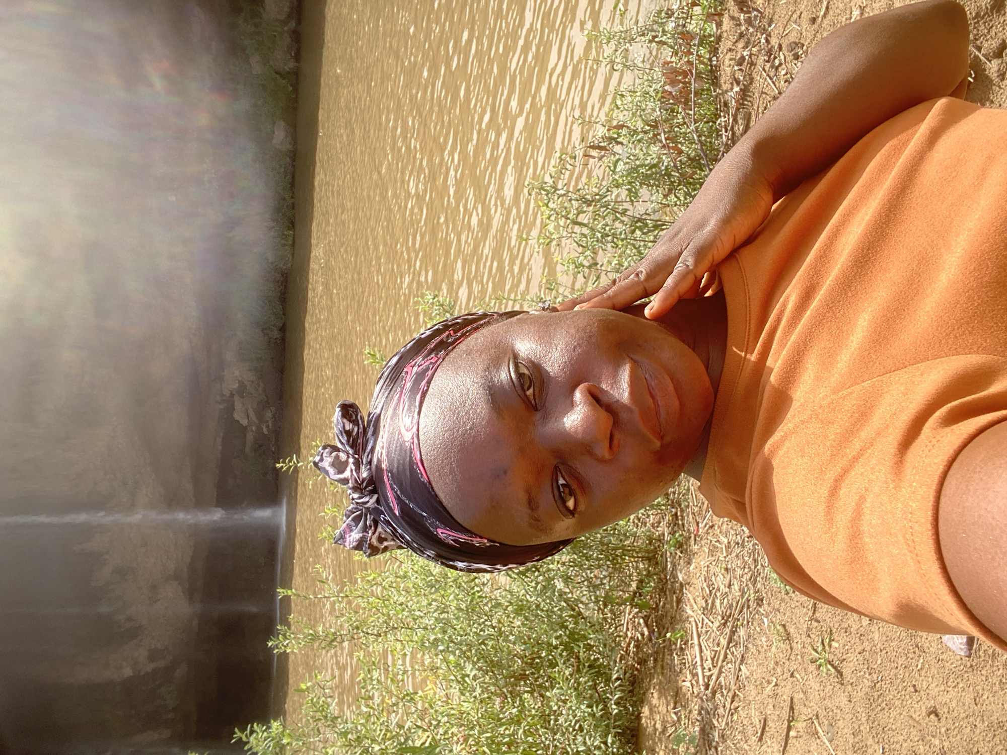 A young woman is captured in a selfie-style photograph, positioned in the foreground of an outdoor scene, likely in Kukum, Nigeria. She has dark skin and is looking directly at the camera with a neutral to slight smile. She wears an orange t-shirt and a patterned headscarf, predominantly black with pink and brown details. Her right hand is gently placed near her cheek.
Behind her, a wide body of water, brownish in color, stretches across the middle ground, reflecting the bright daylight. The immediate foreground consists of dry, sandy earth with patches of green, low-lying vegetation. In the distant background to the left, a waterfall or water cascade is visible, descending from a lighter-colored cliff into the main body of water.
The scene is brightly lit by the sun, indicating it is daytime. A prominent lens flare, appearing as a rainbow-colored haze, is present on the left side of the image, partially overlapping the waterfall. No text is visible.