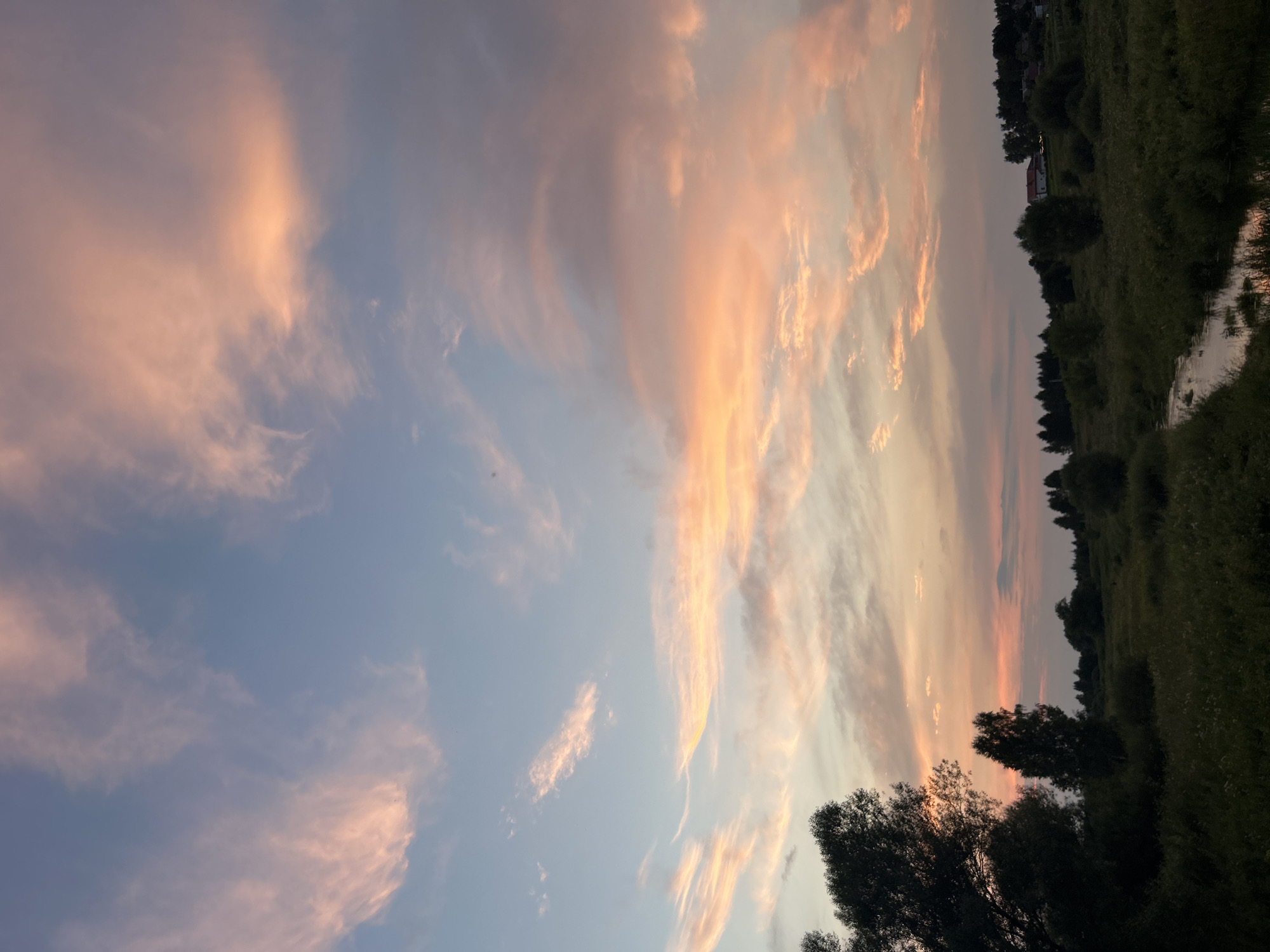 A serene, wide-angle shot captures the natural beauty of a sunset over a rural landscape in Kalitsino, Russia. The sky is a painterly blend of soft blues and vibrant oranges, with wispy clouds tinged with pink and peach hues, illuminated by the setting sun. In the foreground, lush green vegetation blankets the rolling hills, with a small, gentle stream meandering through the scene, its surface reflecting the warm sky. Dark silhouettes of trees line the horizon, interspersed with a few scattered buildings, hinting at a small settlement. The overall atmosphere is one of tranquility and natural splendor.