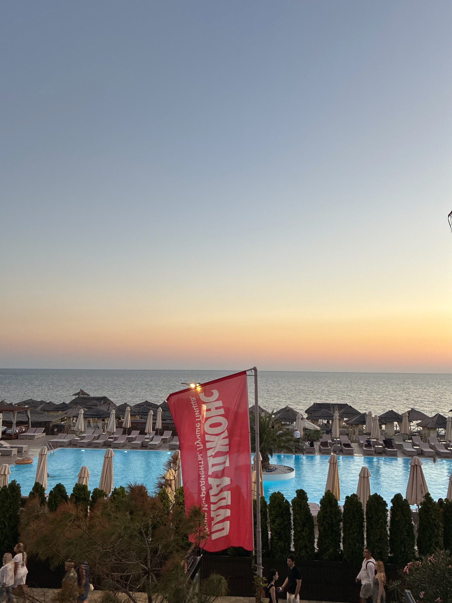 A resort area by the Black Sea in Sochi, Russia, is depicted at sunset. The foreground features a vibrant red flag with white lettering, partially obscuring a swimming pool. The pool is illuminated and surrounded by lounge chairs and umbrellas, suggesting a lively atmosphere. Beyond the pool, thatched-roof cabanas line the beach, with more umbrellas scattered across the sand. The sea is calm and stretches to the horizon, where the sky displays a gradient of soft orange and blue hues, characteristic of a sunset. Several people are visible in the foreground and near the pool, some walking, others relaxing. The overall scene conveys a sense of leisure and vacation.