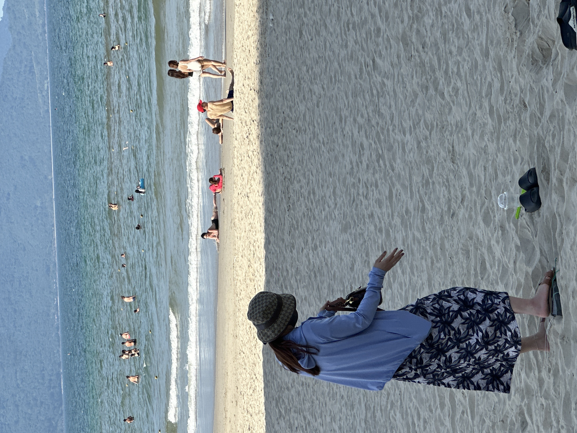 The image shows a busy beach scene at My Khe Beach, Đà Nẵng, Vietnam. In the foreground, a woman wearing a light blue hoodie, a patterned wide-brimmed hat, and a floral skirt stands on the sand. She is holding a dark handbag and has her right arm extended, as if gesturing. Her feet are bare, wearing simple flip-flops. Scattered on the sand near her are a pair of black flip-flops, a clear plastic cup, and a green object.
In the midground, several people are visible on the sandy beach and in the shallow water. Some individuals are sunbathing, including a person wearing a red shirt and another in black swim trunks. Two women in swimsuits are walking towards the water.
The background features the turquoise waters of the sea, with numerous people swimming and playing. Yellow buoys are scattered in the water, marking off areas. The distant horizon is dominated by a hazy, green mountain range, characteristic of the Đà Nẵng coastline. The sky is clear and bright, indicating a sunny day. The overall scene depicts a lively recreational atmosphere typical of a popular beach destination.