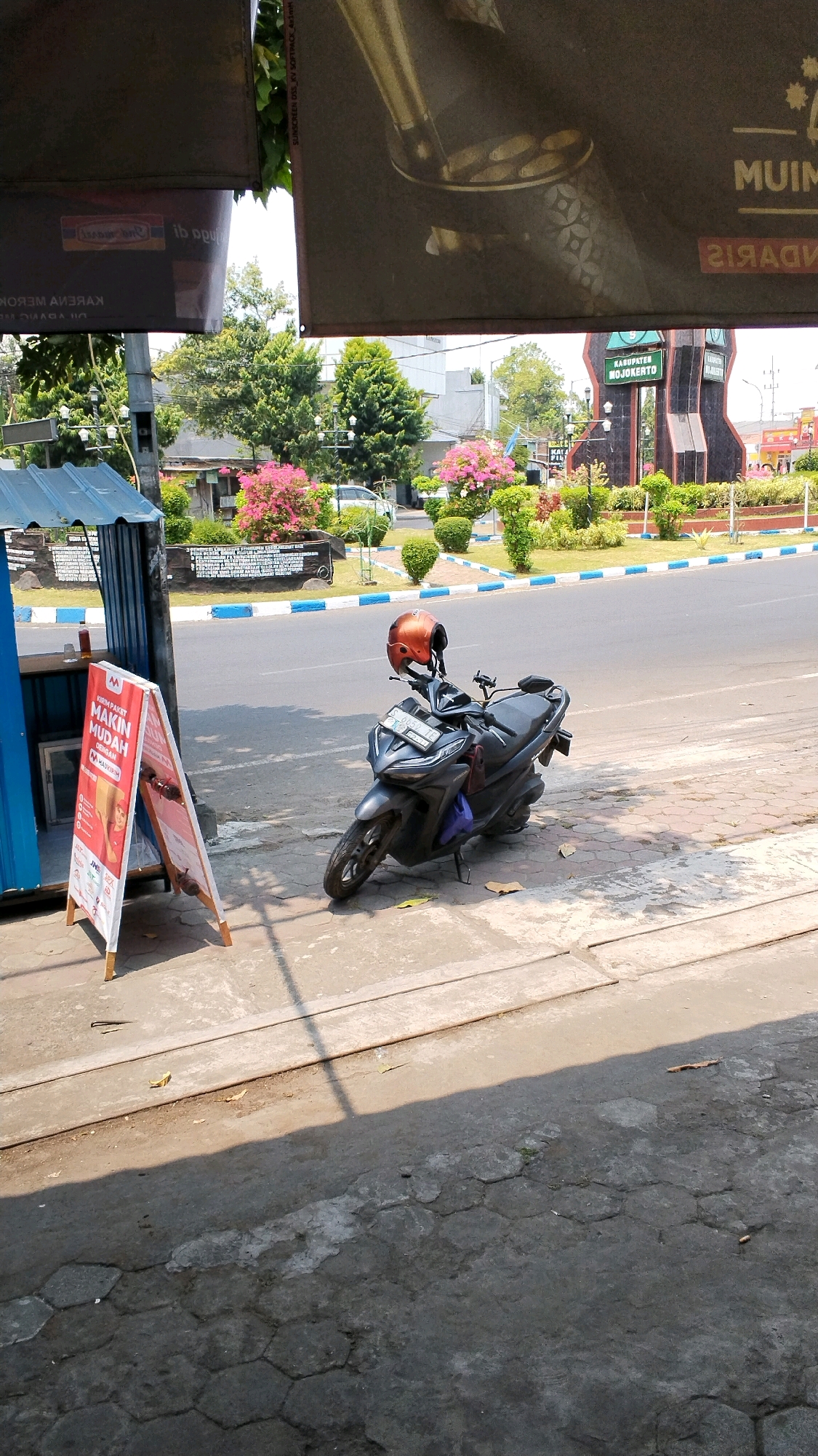The image depicts a street scene in Banjaragung, Indonesia, with a focus on a dark grey motorcycle parked on a sidewalk. The motorcycle has an orange helmet resting on its handlebars. To the left of the motorcycle, a small blue and silver kiosk or stand is visible, with a red and white A-frame sign in front of it. The sign displays text and logos, suggesting it is advertising a service. Behind the kiosk, a white car is partially visible.
The sidewalk is paved with hexagonal concrete blocks and a portion of it is in shadow. A road runs alongside the sidewalk, with a blue and white striped curb. In the background, there are various trees and shrubs, some with pink flowers, and a prominent monument or landmark with the text "KABUPATEN MOJOKERTO" visible. Overhead, there are dark tarpaulins or banners. The scene is illuminated by bright sunlight, casting shadows on the ground. The overall impression is of a typical day on a street in a populated area.