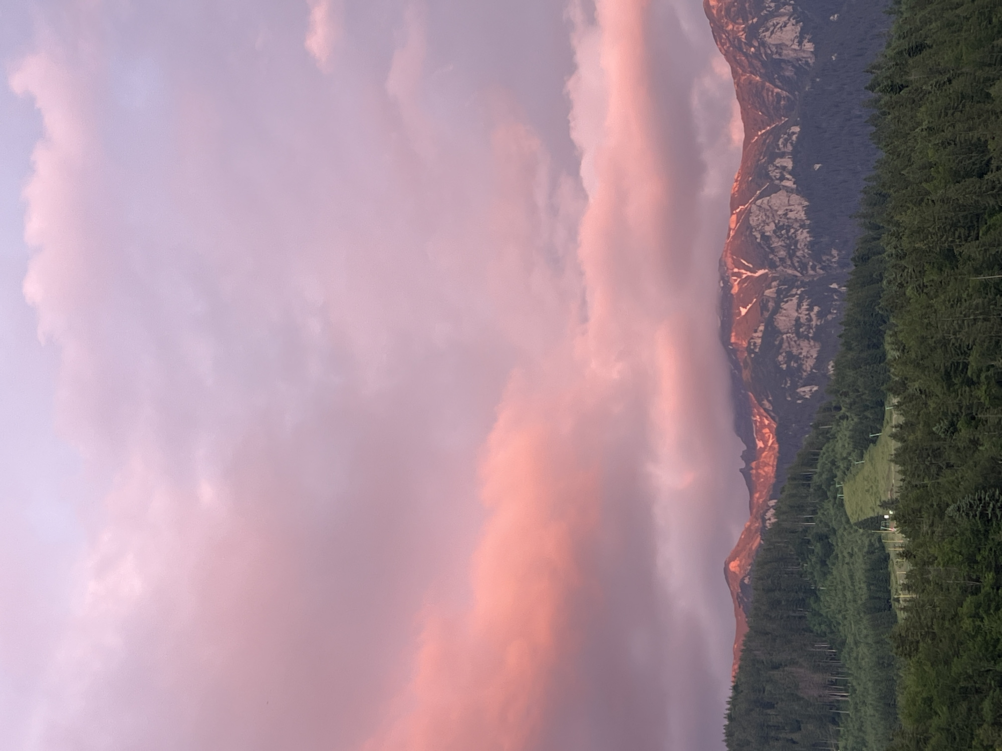 An aerial shot captures a majestic mountain range bathed in the soft, pink hues of a sunset. The sky is a canvas of pastel colors, with streaks of vibrant pink and soft lavender clouds. The mountain peaks, jagged and imposing, are illuminated by the setting sun, casting a warm glow on their rocky surfaces. Patches of snow are visible on the higher elevations, contrasting with the dark, dense evergreen forests that blanket the lower slopes. The trees form a rich, textured carpet that stretches across the landscape, creating a dramatic backdrop for the illuminated peaks. The scene is one of natural beauty and tranquility, evoking a sense of awe and wonder.