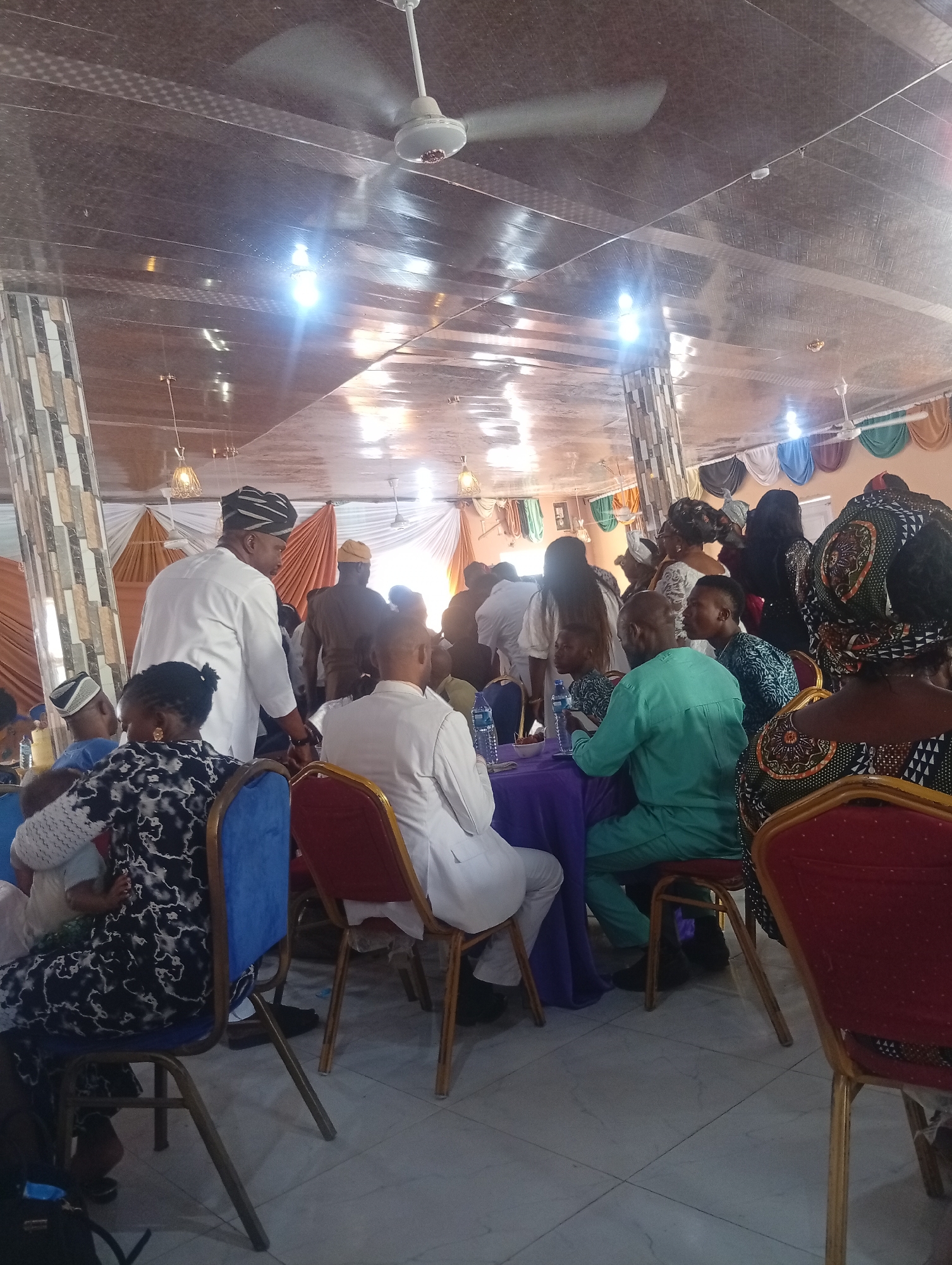 The image captures an indoor gathering in Dutse, Nigeria, likely a celebratory event or meeting, based on the attire and ambiance. The room has a high ceiling with a reflective surface, adorned with ceiling fans and bright fluorescent lights, some of which are visible as glare. Decorative curtains in various colors—brown, orange, white, green, blue, and red—are hung along the walls. The floor appears to be tiled with a light grey grid pattern.
In the foreground, several individuals are seated. A woman in a black and white patterned dress is visible from the side, seated on a blue chair. To her right, a man in a white traditional outfit is facing away, and another man in a light green traditional outfit is seated at a table covered with a purple cloth, interacting with someone across the table. A partially visible person in a red chair is also present.
Further back, more people are gathered, some standing and some seated. A man in a white outfit with a patterned black and white cap is a prominent figure in the mid-ground, seemingly interacting with others. Other individuals in traditional Nigerian attire are dispersed throughout the scene. The overall atmosphere suggests a social or formal occasion with a sense of community and festivity.
