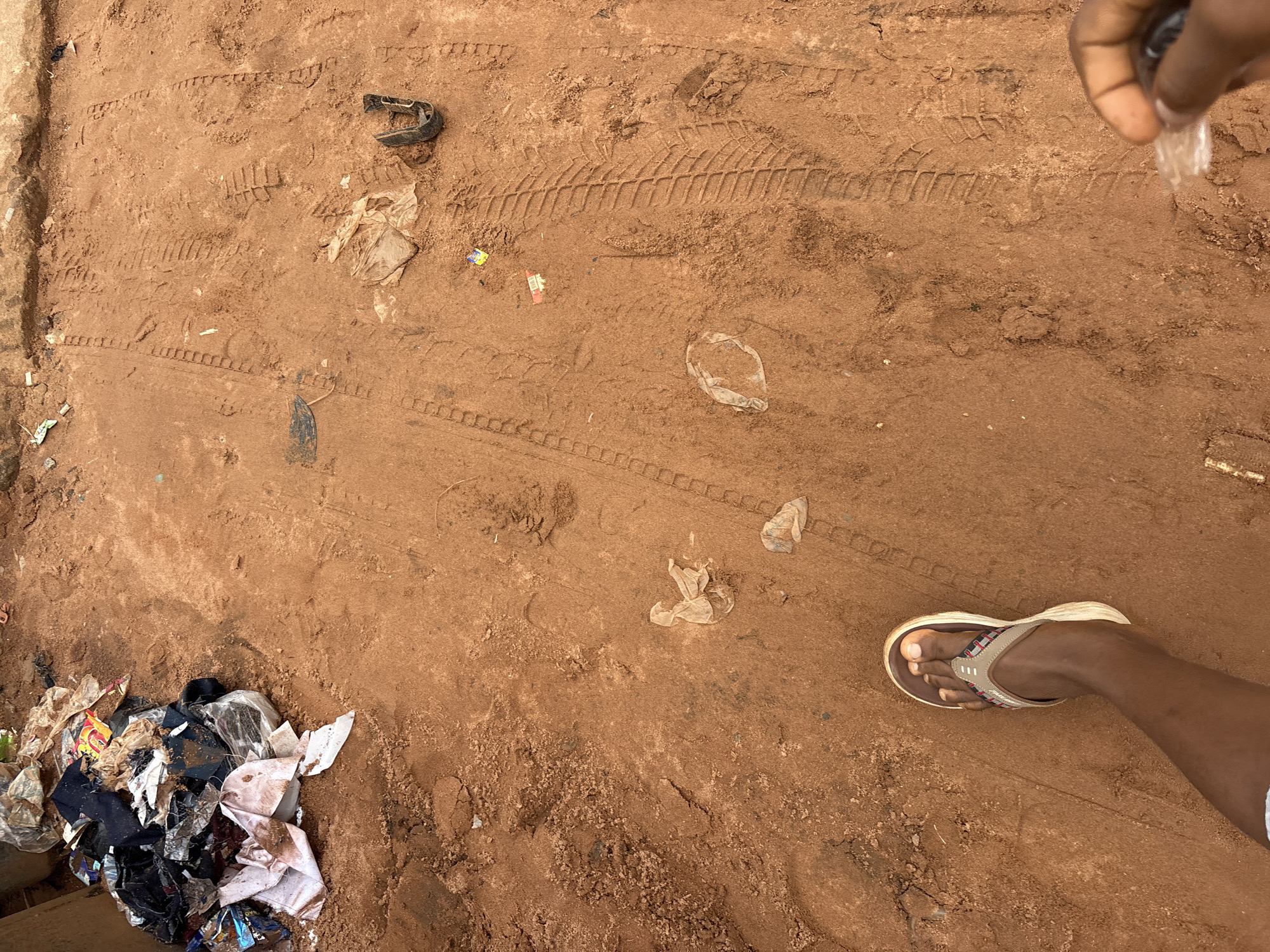 This image depicts a close-up view of a dirt ground surface, likely in the city of Nkpor, Nigeria. The ground is reddish-brown and shows distinct tire tracks from a vehicle, as well as various scattered pieces of trash. Among the debris, one can observe torn clothing, plastic bags, and other unidentifiable refuse. In the lower left corner, a person's left foot, clad in a flip-flop, is visible, suggesting human presence. To the right, a hand is holding a small, clear plastic object. The overall scene conveys a sense of neglect and an unhygienic environment, possibly an area where waste has accumulated.