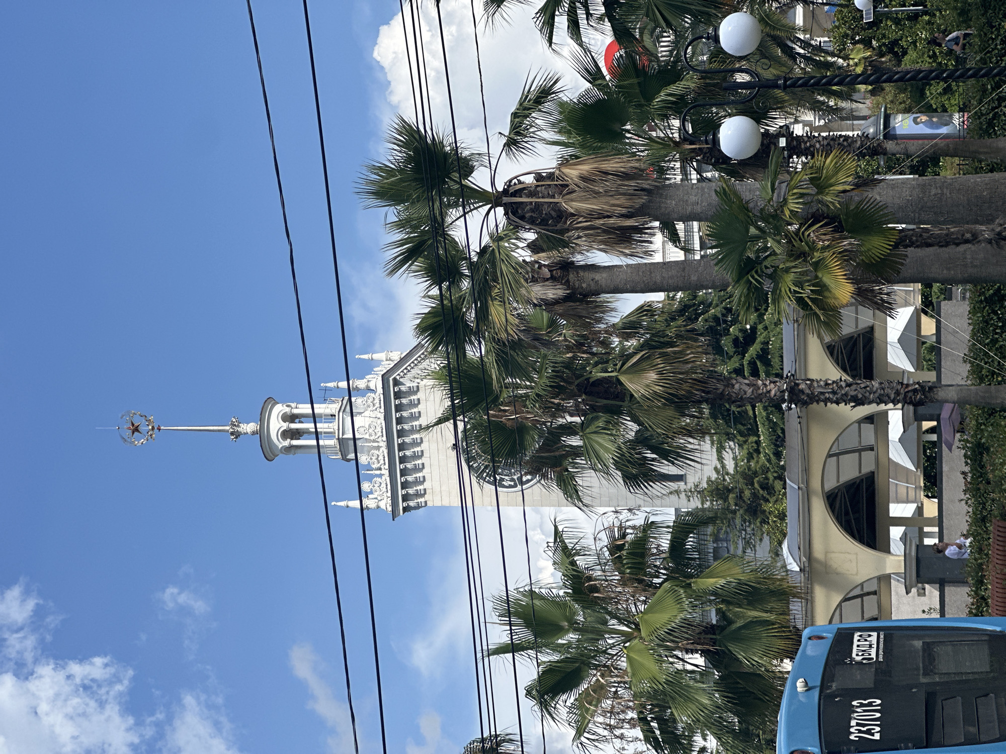 The image shows a view of the Sochi Sea Port building in Sochi, Russia, on a sunny day with a partly cloudy sky. The prominent white building features a tall tower topped with a red star and a decorative spire. A large clock face is visible on the tower. In the foreground, several palm trees of varying sizes are present, their fronds casting shadows. Overhead, black electrical wires crisscross the frame. A blue bus with the number "237013" and Russian text is partially visible on the left side of the image. An archway structure is visible behind the palm trees. A person in a white shirt is standing near a low wall. Two ornate lampposts with globe-shaped lights are also visible, along with a poster or advertisement. The scene appears to be a public area, possibly a waterfront promenade.