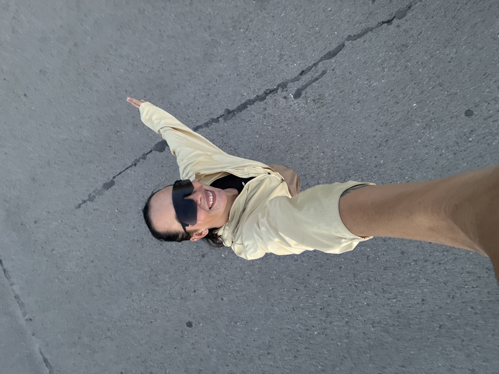 A selfie taken from a high angle shows a person standing on an asphalt surface, possibly a road or sidewalk, in Chaikovsky, Russia. The person is wearing sunglasses and a light-colored jacket, with their right arm extended, holding the camera. Their left arm is also extended, with fingers spread open. The background is a gray, textured asphalt with faint cracks and a dark line running diagonally across the frame. The lighting suggests it is daytime, possibly late afternoon or early morning due to the soft light.