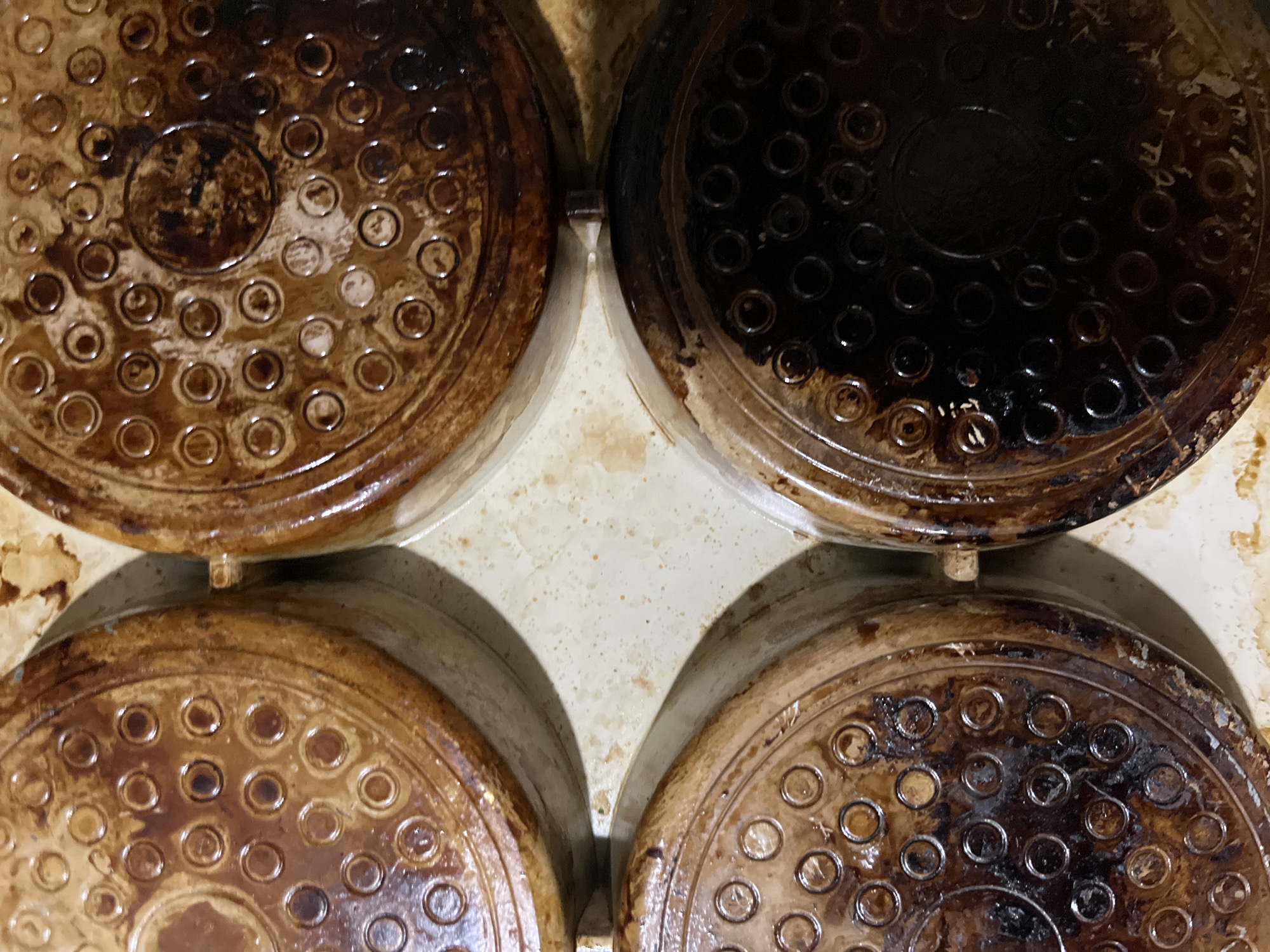 The image shows the bottoms of four waffle maker plates, each with a pattern of circular indentations. The plates are positioned in a square arrangement, with two on the top row and two on the bottom row. They appear to be made of a metal material and are heavily soiled with dark, burnt-on residue, suggesting they have been used extensively. The residue varies in color from dark brown to black, with some lighter, greasy smudges visible as well. The waffle maker itself appears to be made of a light-colored, possibly ceramic or plastic material, which is also stained in places. The overall impression is one of a well-used kitchen appliance that is in need of cleaning. There is no visible text in the image. The lighting is somewhat harsh, casting shadows and highlighting the texture of the burnt-on food. The image is taken from a top-down perspective, focusing on the waffle plates. No people are visible. The setting appears to be a kitchen or storage area for kitchen equipment.