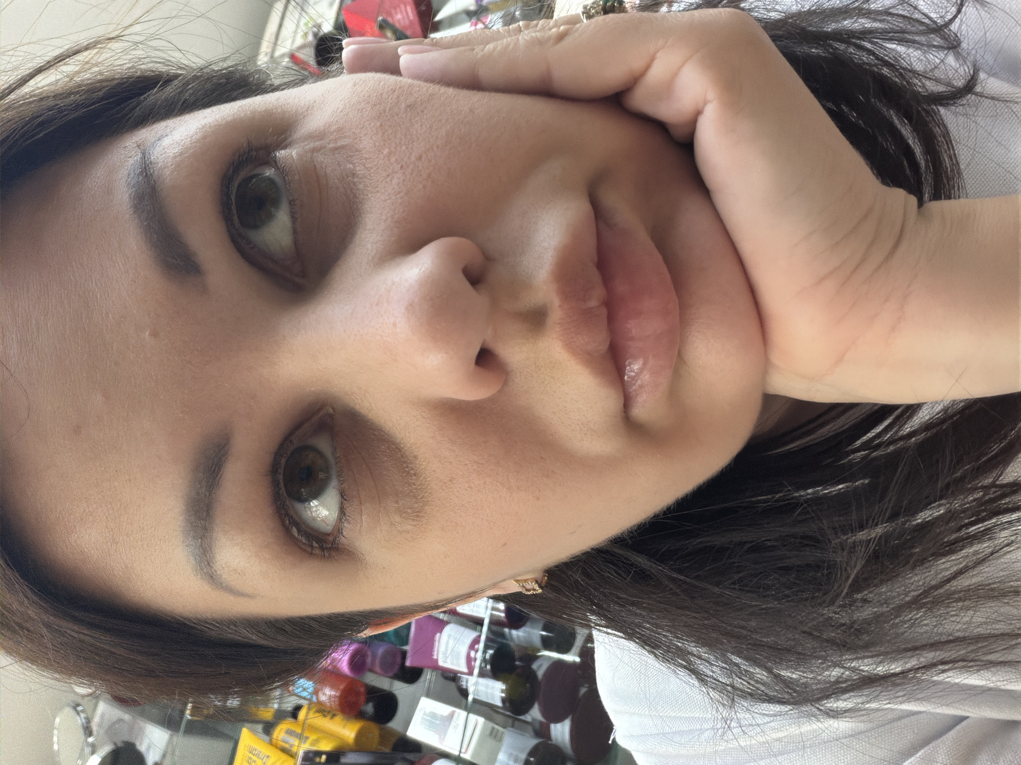 The image is a close-up, head-and-shoulders portrait of a woman with dark hair, resting her chin on her right hand. She is wearing a white top and a small gold earring. Her eyes are looking upwards, and her lips are slightly parted. The background shows shelves stocked with various products, appearing to be a retail setting. There is no information to indicate this scene is located in the city of Sochi, Russia.
