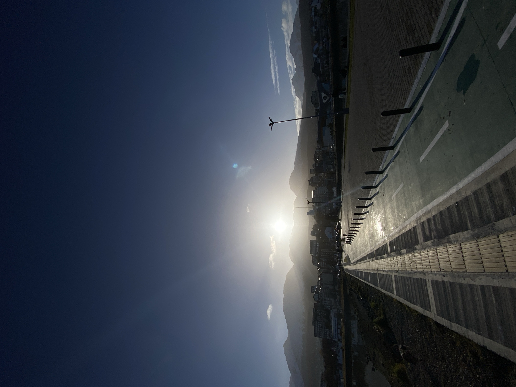 The image depicts a landscape view of Ushuaia, Argentina, taken at a low angle against a bright sun. The foreground features a paved walkway with a green-painted bike lane marked by white lines. Dark posts are positioned along the edge of the walkway.
In the middle ground, a city sprawls at the base of mountains. Buildings are discernible, their details obscured by the bright sun. A tall, thin structure resembling a windmill stands near the city's edge.
The sky is a gradient of deep blue to light blue, and the sun is shining brightly, creating a strong glare. The overall impression is of a bright, sunny day in a city situated in a mountainous region.