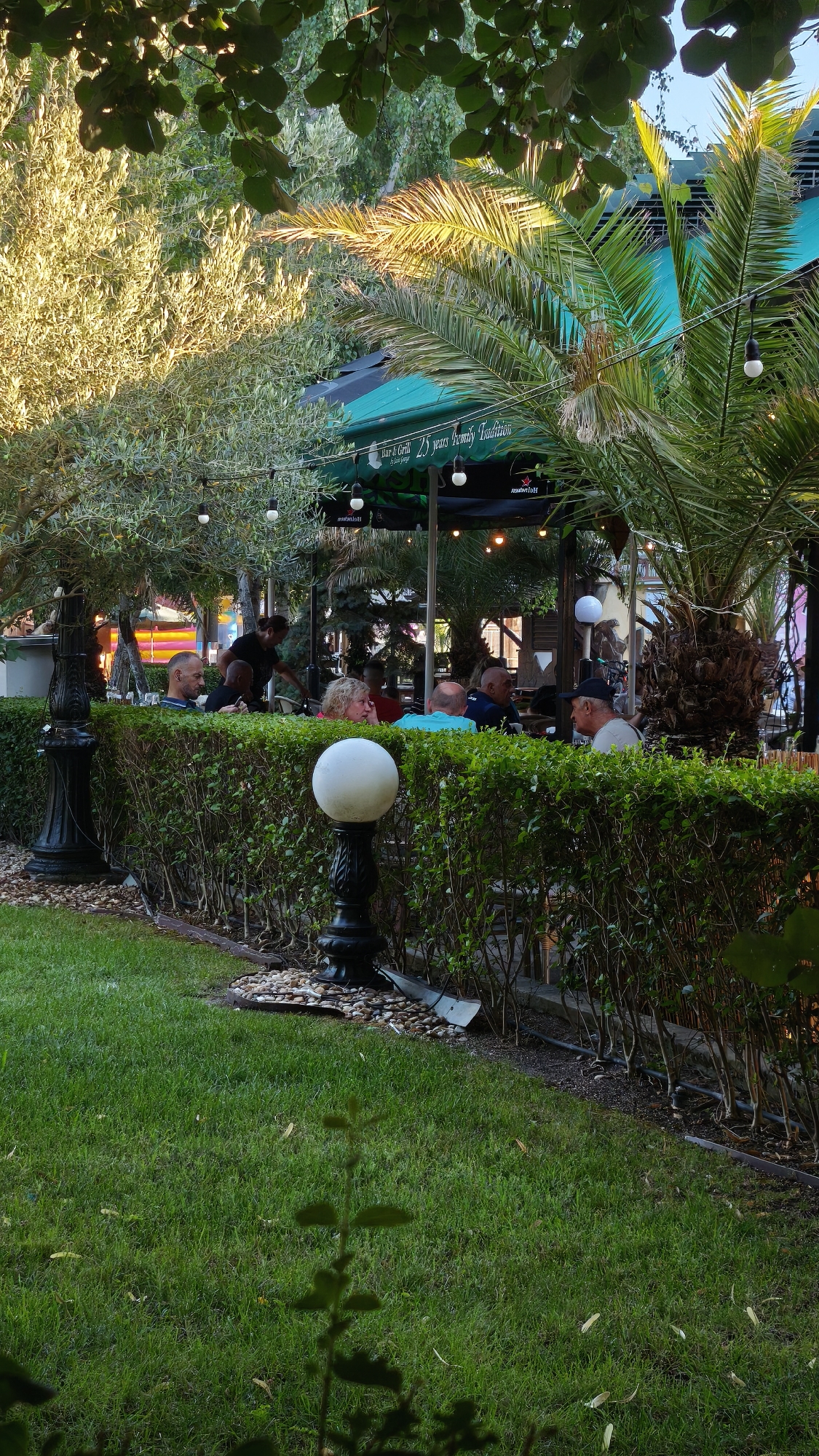 The image depicts an outdoor seating area of a restaurant, likely in the city of Pomorie, Bulgaria, judging by the context. The scene is set during daylight, with sunlight filtering through lush greenery, including trees and palm fronds, suggesting a warm climate. In the foreground, a well-maintained lawn is visible, with a decorative hedge running horizontally across the frame. Along the hedge, a black, ornate lamp post with a white spherical light fixture stands. Beyond the hedge, several people are seated at tables under green umbrellas, indicating a dining or social gathering. One person, seemingly a waiter, is standing and attending to a table. The atmosphere appears relaxed and leisurely, characteristic of a restaurant's outdoor terrace.