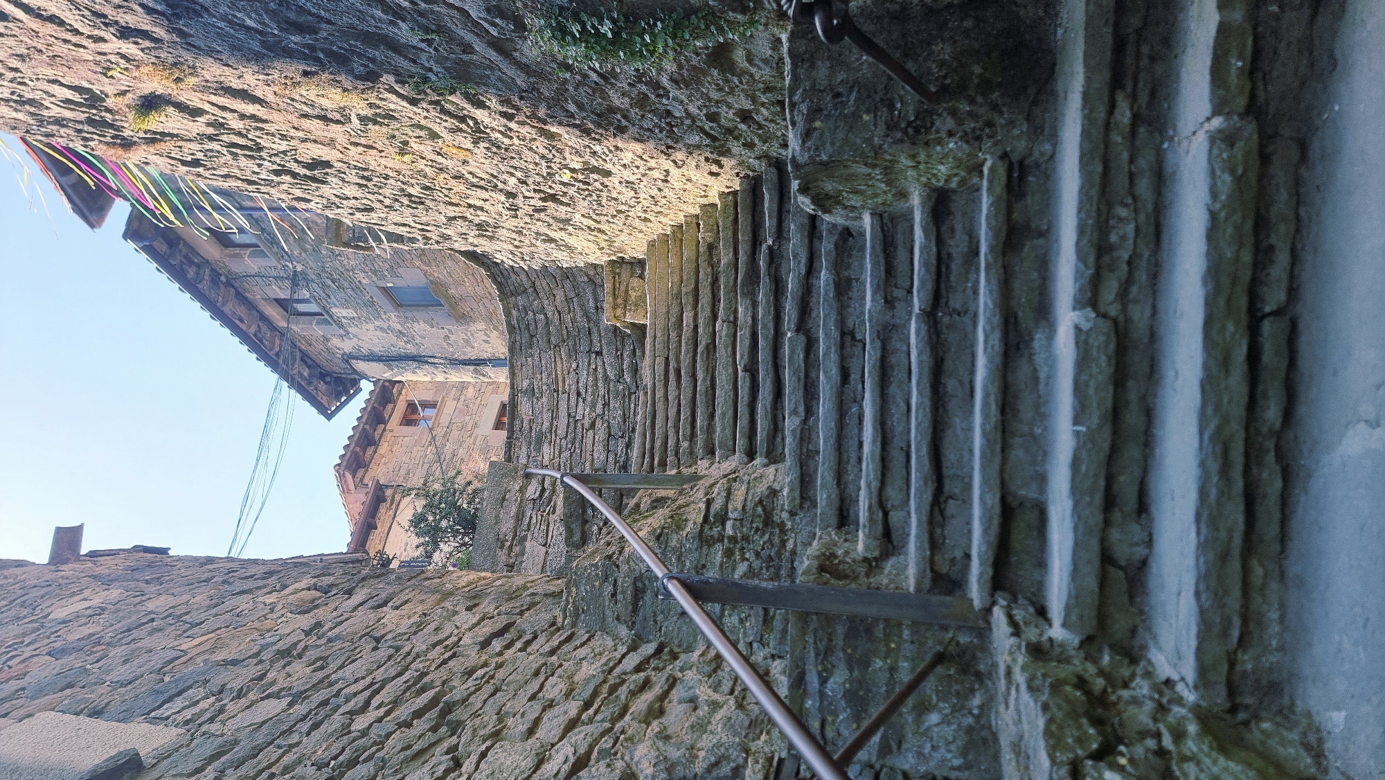 This is a photograph of an old stone staircase in the medieval village of Rupit i Pruit, Catalonia, Spain. The staircase is made of uneven, weathered stone steps and is flanked by rustic stone buildings. A metal handrail runs along the left side of the stairs. The buildings have stone walls and tile roofs, and the overall impression is one of antiquity and a historic European setting. The sky is visible at the top of the image, and there are some overhead wires and colorful ribbons strung between buildings. The lighting suggests it is daytime, with sunlight casting shadows on the scene.