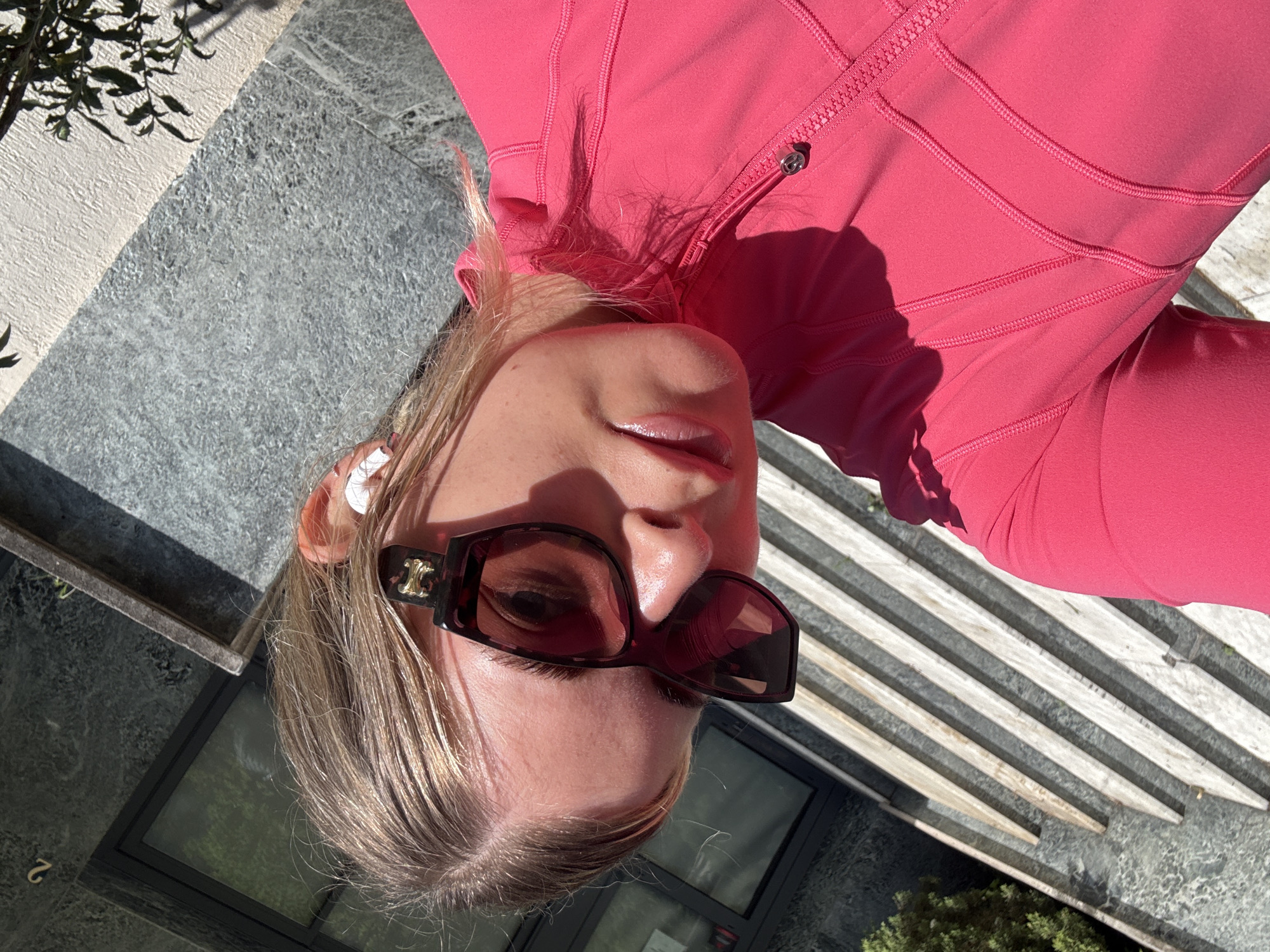 A blonde woman wearing dark-framed sunglasses is lying on stone steps. She is wearing a pink zippered top. The steps are stone with a pattern of dark and light grey. Behind the steps is a dark surface that reflects trees and greenery. The location appears to be outdoors in a location with architectural elements consistent with Paris, France.