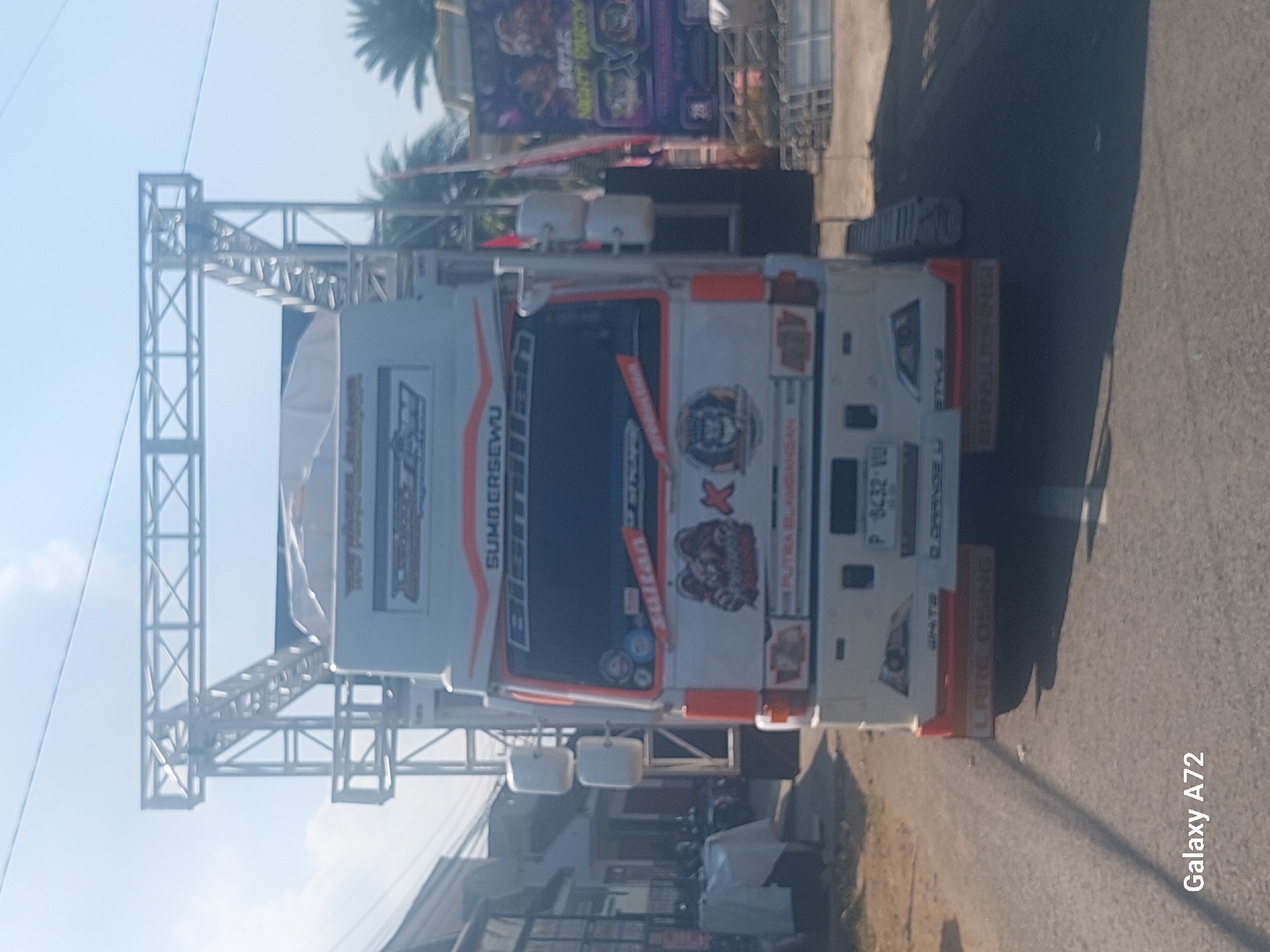 A white truck with an elaborate sound system mounted on top is parked on a street in Sengguruh, Indonesia. The truck is decorated with various decals, including a large image of a lion and the words "SUMBERSEWU" and "PUTRA BLANGSAK." The truck's license plate reads "P 8432 VU." The background shows a row of buildings, palm trees, and a clear blue sky. The image appears to have been taken during the daytime.