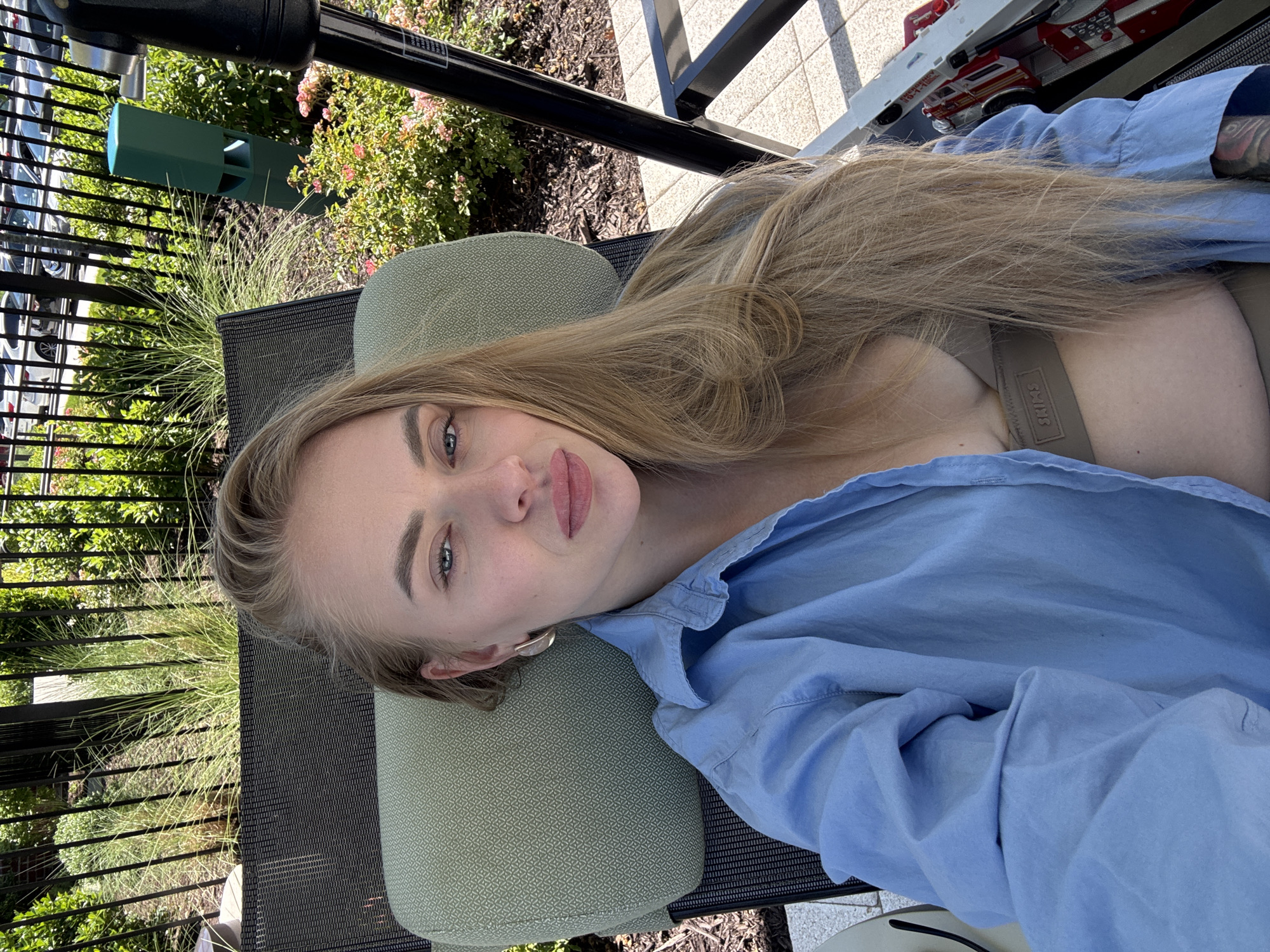 A fair-skinned woman with long, light brown hair is lying on an outdoor chaise lounge. She is wearing a light blue button-up shirt over a tan sports bra. Her head is resting on a patterned green pillow. The chaise lounge is black metal with a mesh seat. Behind the lounge is a black metal fence, a green structure, and various plants. To the side of the woman are tiled squares, upon which sits a toy fire truck. The location is unknown.
