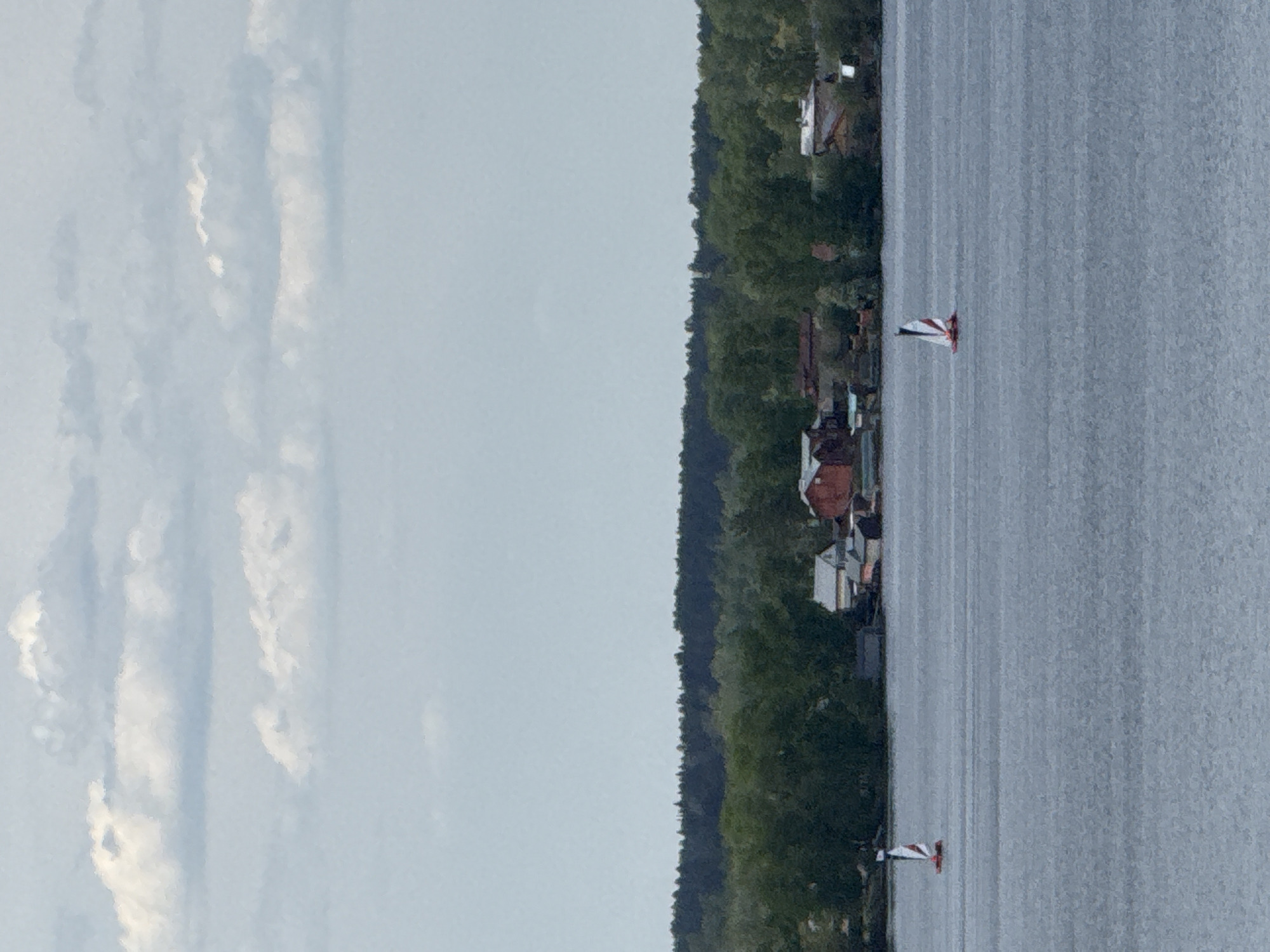 The image depicts a body of water, likely a lake or wide river, with trees lining the shore. Several buildings are visible among the trees. Two sailboats with white and red sails are present on the water. The sky is overcast with long, streaky clouds. The location is potentially in Votkinsk, Russia.