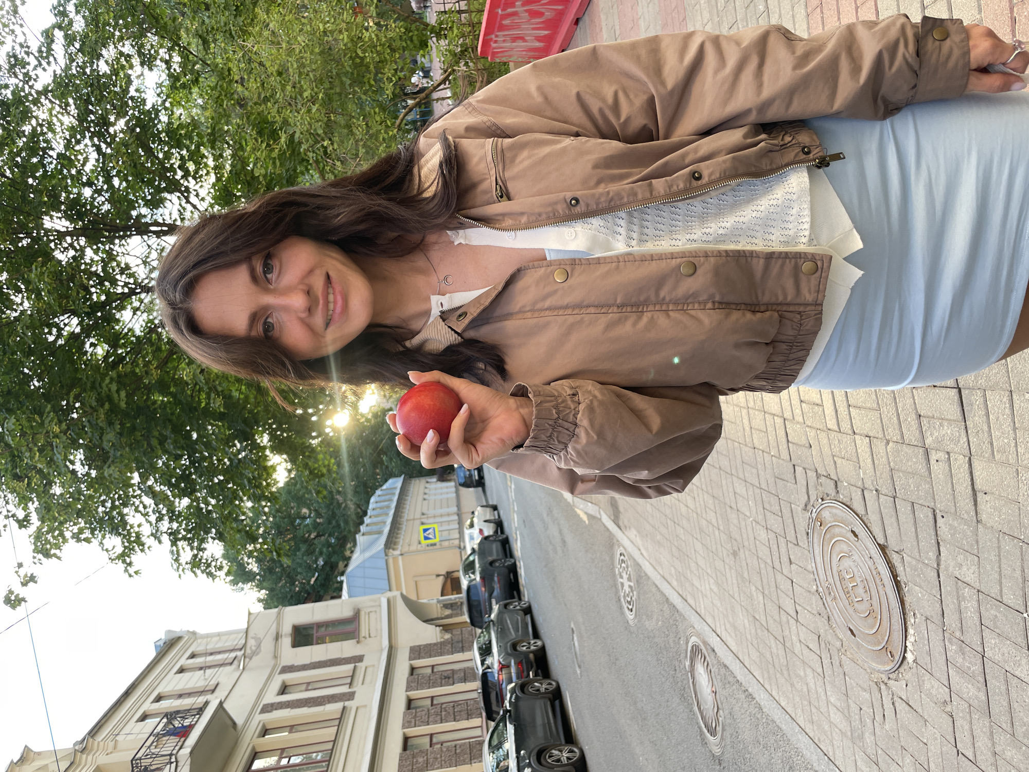 A young woman stands on a sidewalk in Saint Petersburg, Russia, holding a red apple. She has long, dark brown hair and is wearing a light brown bomber jacket over a white top and a light blue skirt. The sun is setting behind her, casting a warm glow on the scene. Cars are parked along the street to her left, and a building with architectural details typical of Saint Petersburg is visible behind them. The sidewalk is paved with bricks, and manhole covers are visible on the ground. A red barrier with graffiti is present in the background.