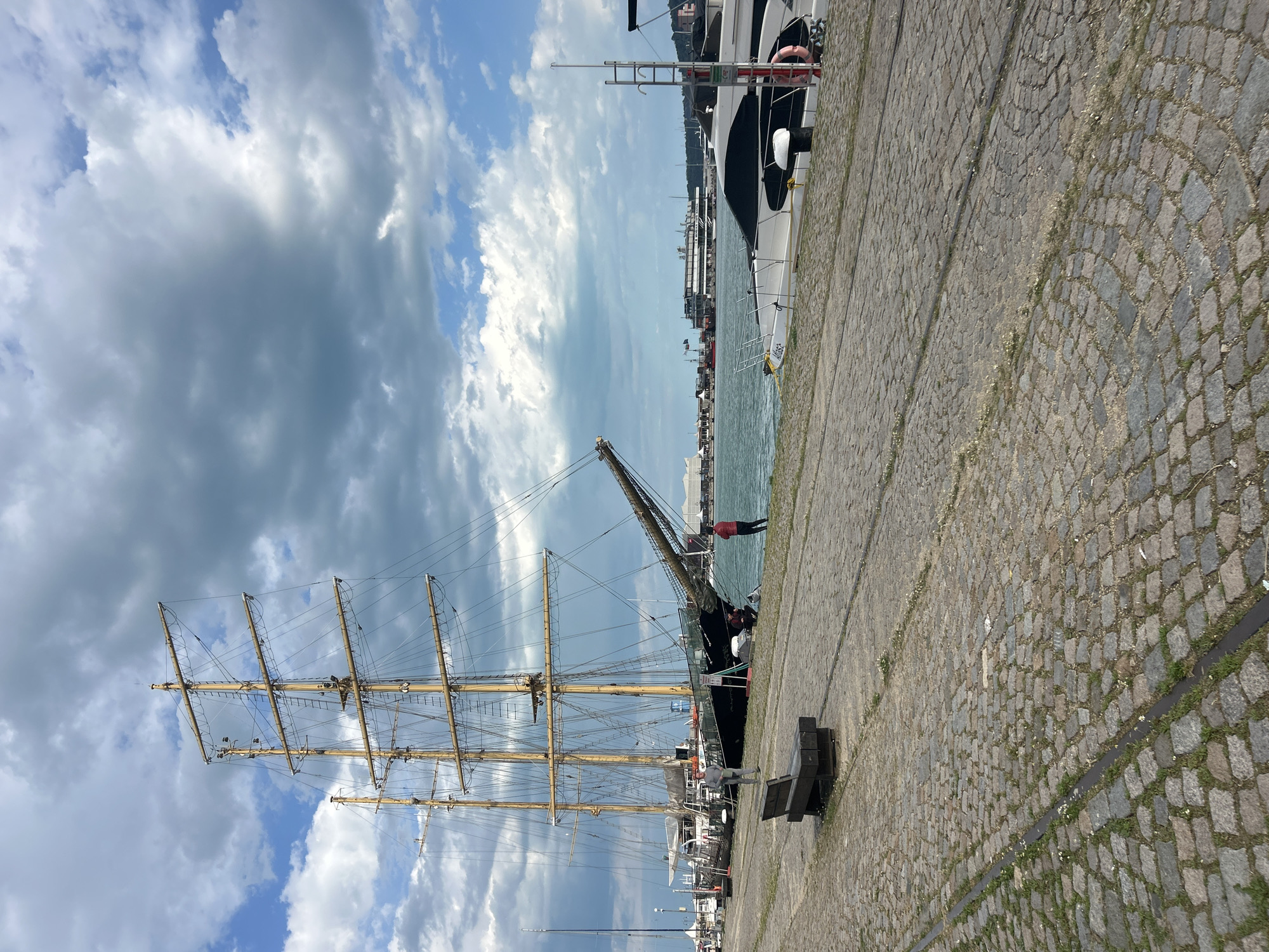 The image shows a tall sailing ship docked in Varna, Bulgaria. The ship has multiple masts and rigging extending upwards against a sky with a mix of blue patches and heavy, grey clouds. A crane is attached to the ship's side.
The ship is docked along a quay paved with cobblestones. In the background, other boats and city buildings are visible across the water. A person in red stands near the edge of the quay, while another person in gray is seen near the ship. A bench is situated along the quay.