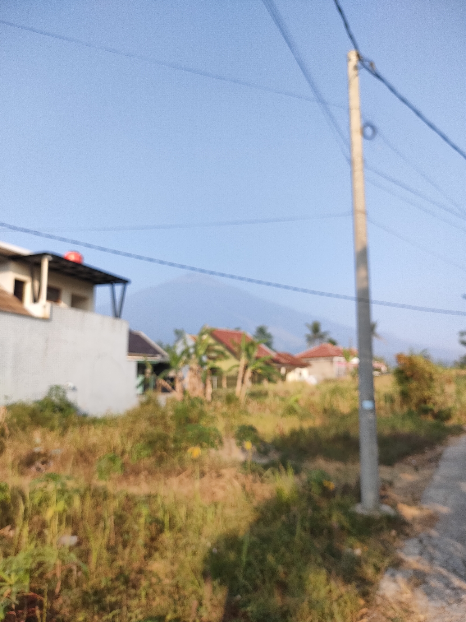 The image depicts a blurry outdoor scene with a clear sky and a distant hazy mountain. In the foreground, a utility pole stands tall, with power lines crossing the frame. To the left, a portion of a light-colored building with a dark roof and some greenery is visible. Further back, several houses with red roofs are scattered amongst dry grass and trees. A paved path runs along the right side of the image. The overall impression is of a rural or suburban area. The location is identified as Caracas, Indonesia.