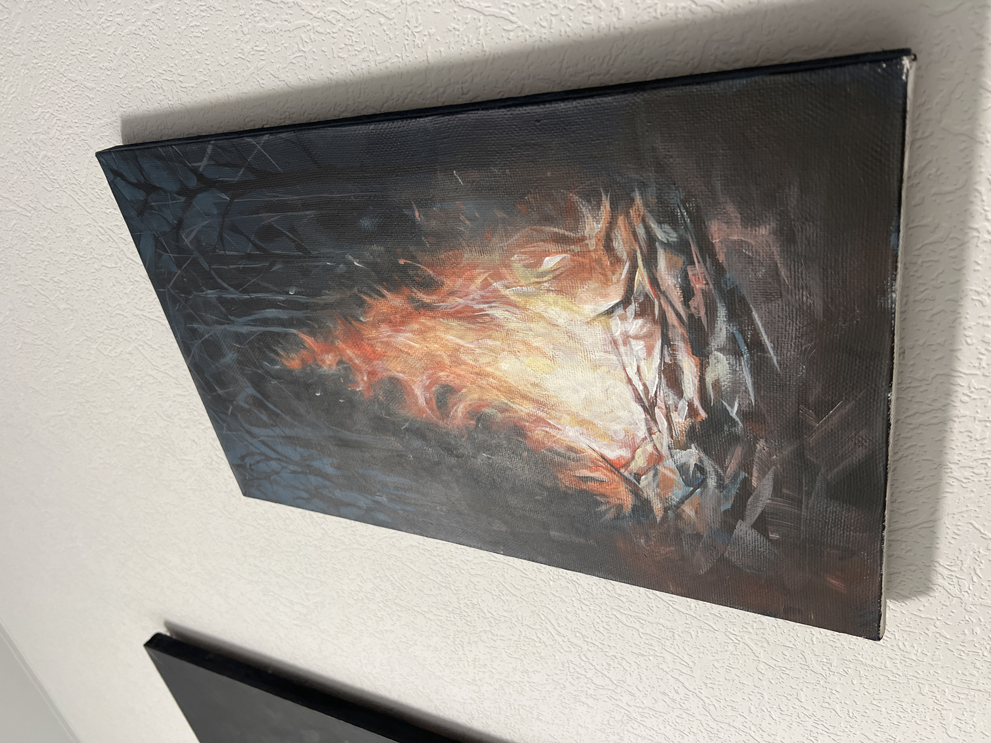 The image shows an oil painting hanging on a white textured wall. The painting depicts a dark, abstract scene with orange and white highlights in the center, possibly representing fire or light. The edges of the painting are black, and the canvas is stretched over a wooden frame. A small portion of another, darker painting is visible below the main painting. The photograph is taken indoors under artificial lighting. The location in Tyumen, Russia is not specified in the image.