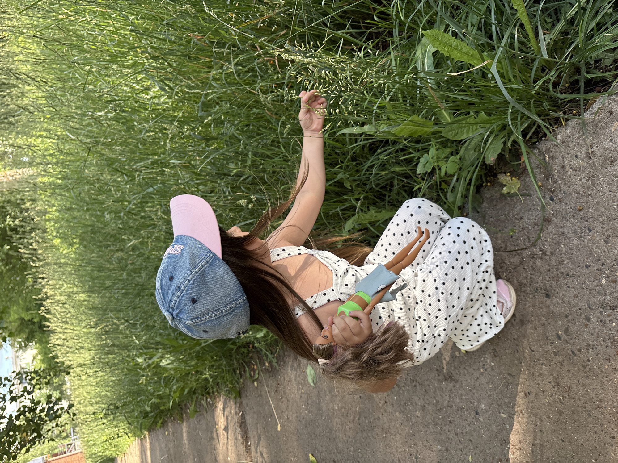 In Chaikovsky, Russia, a young girl wearing a denim baseball cap and a white polka-dot dress is crouched on a paved path next to a tall, green grassy area. She is holding a doll in her left hand and reaching out with her right hand to touch the grass. The lighting suggests it is daytime, with sunlight illuminating the scene. The environment appears to be an outdoor urban setting, possibly a park or a residential area with green spaces.