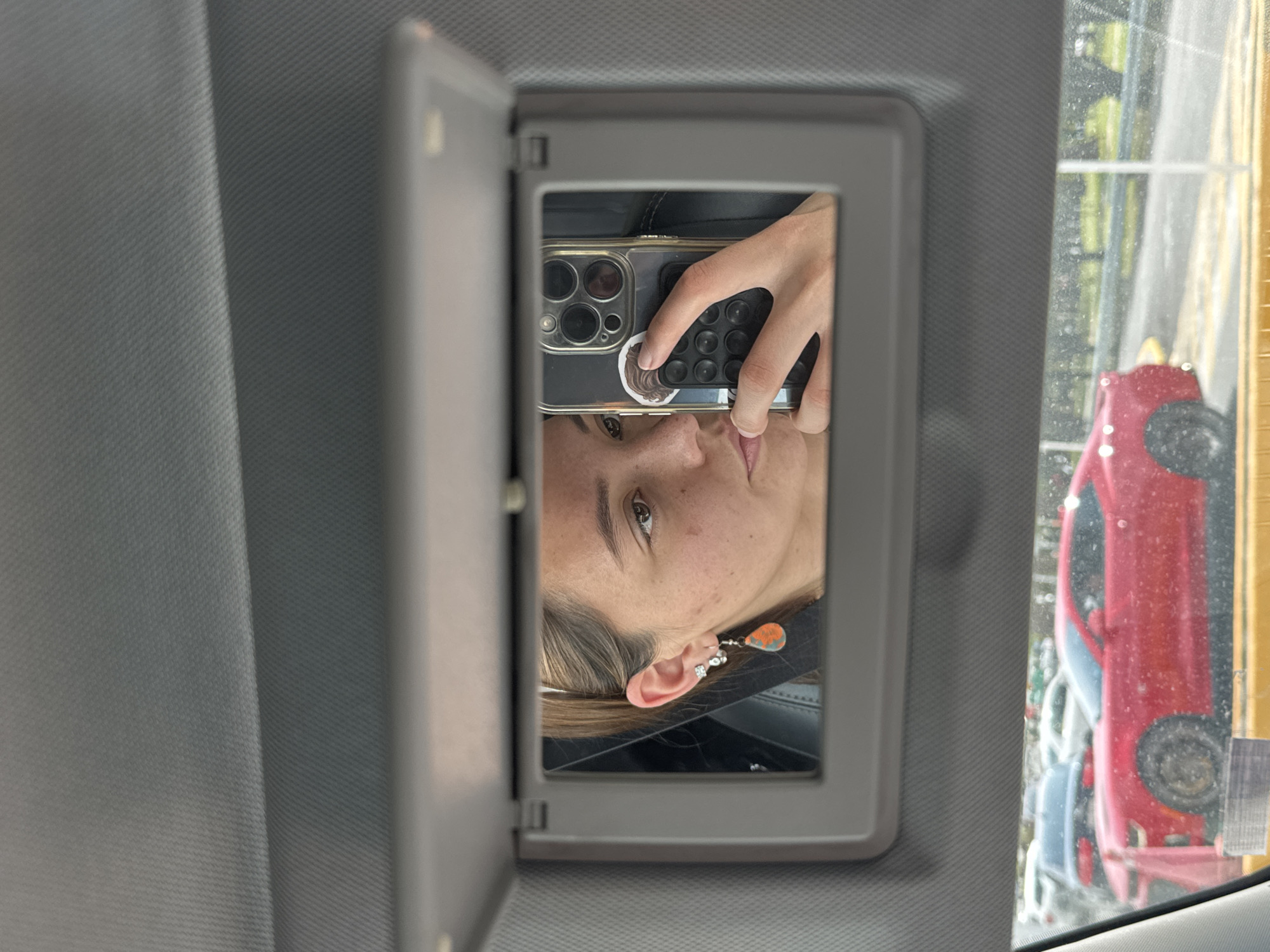 A young woman is taking a selfie in the mirror of a car's sun visor. She is holding a black iPhone with a gold case. Her face is partially visible, with a focus on her eye and ear, where she wears multiple earrings. Outside the car window, there are cars parked. Based on the surroundings, the location is determined to be in San Andrés Cholula, Mexico.