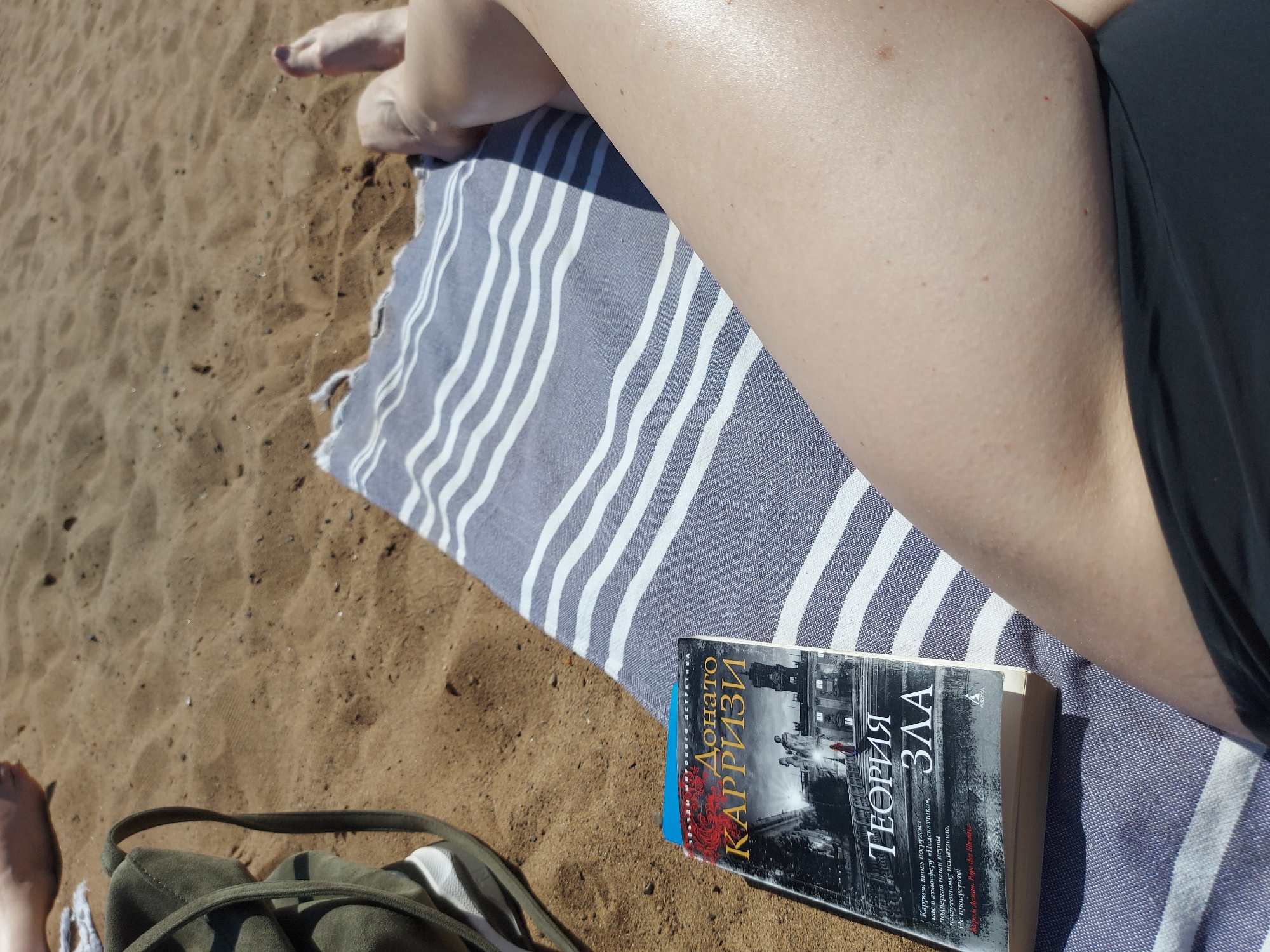 The image displays a high-angle shot of a person relaxing on a sandy beach or riverbank, consistent with the outdoor environment found near Chaikovsky, Russia. The scene suggests a leisurely activity taking place during bright daylight.
The main subjects are the person's legs, a striped beach towel, and a book. The person's bare legs, including their upper thighs and feet, are visible. One foot in the top left corner shows toenails painted a dark color. The skin on the legs appears smooth and slightly reflective from the sunlight. A black swimsuit bottom is visible along the right edge of the frame. The person is lying on a blue and white striped towel, which is spread over the sand.
A book, titled "ТЕОРИЯ ЗЛА" (Theory of Evil) by Donato КАРРИЗИ (Donato Carrisi), is prominently placed on the sand next to the towel. The book's cover features a stark black and white image of buildings and figures, with the author's name and part of the title highlighted in red Cyrillic text. The OCR reveals a blurb on the cover: "Карризи вновь погружает нас в атмосферу «Подсказчика», подвергая наши нервы внешуточному испытанию. Не пропустите! Жером Дежан. Раage des libraires" (Carrisi once again plunges us into the atmosphere of "The Whisperer," subjecting our nerves to a serious test. Don't miss it! Jérôme Dejean. Page des libraires).
The ground is composed of light brown sand, showing visible texture and some small pebbles. A portion of a dark green canvas bag is visible in the bottom left corner, indicating other personal items are nearby. The overall impression is one of peaceful relaxation and reading outdoors on a sunny day.