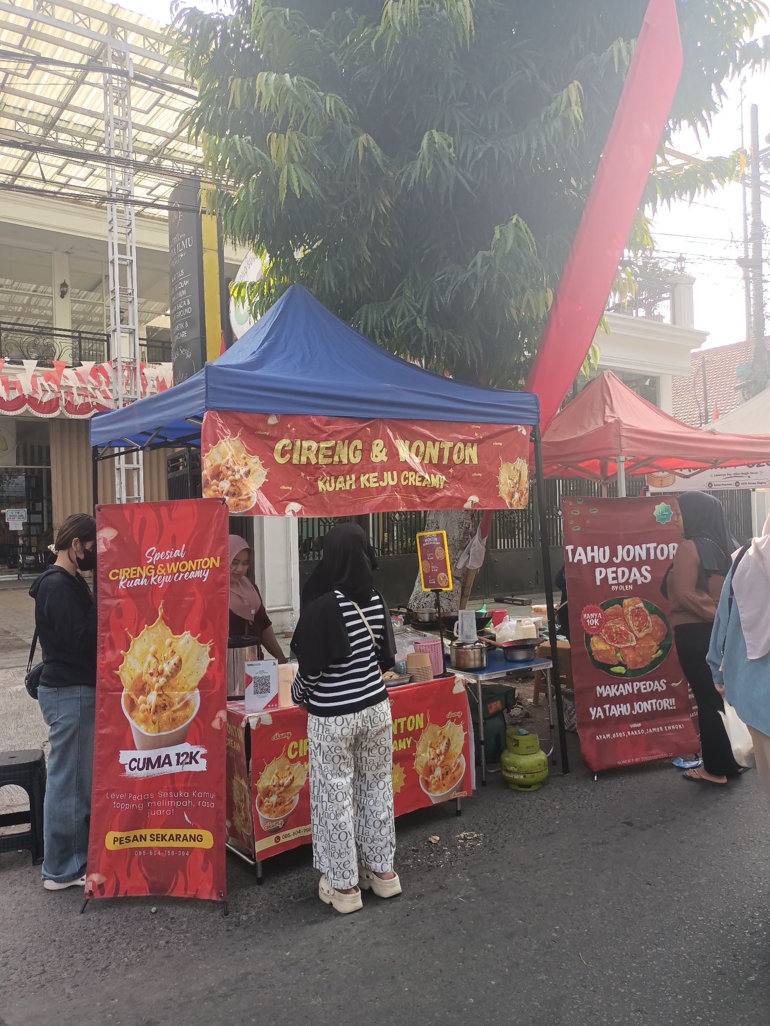 The image shows a street food stall in Pare, Indonesia. The stall is selling cireng and wonton with creamy cheese sauce, as well as spicy tofu. There are two tents, one red and one blue, set up on the side of the street. A woman is standing behind the counter of the stall, serving customers. Two women are standing in front of the stall, looking at the menu. The street is paved and there are trees and buildings in the background. The overall scene is a typical street food market in Indonesia.