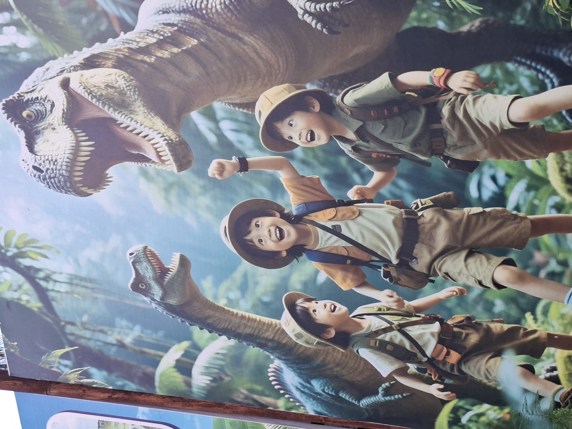 The image is a photograph of a large sign, likely a promotional banner, featuring an illustration of three young boys dressed as safari explorers superimposed in a prehistoric jungle environment with dinosaurs. A large Tyrannosaurus Rex head dominates the upper left corner with its mouth open, while a long-necked Brachiosaurus stretches upward from the bottom of the image. The boys, wearing safari hats and carrying backpacks, appear excited and energetic. The jungle backdrop includes lush green foliage and hints of blue sky.
The photograph seems to have been taken in Lhokseumawe, Indonesia, although this is inferred information not visible in the image itself.