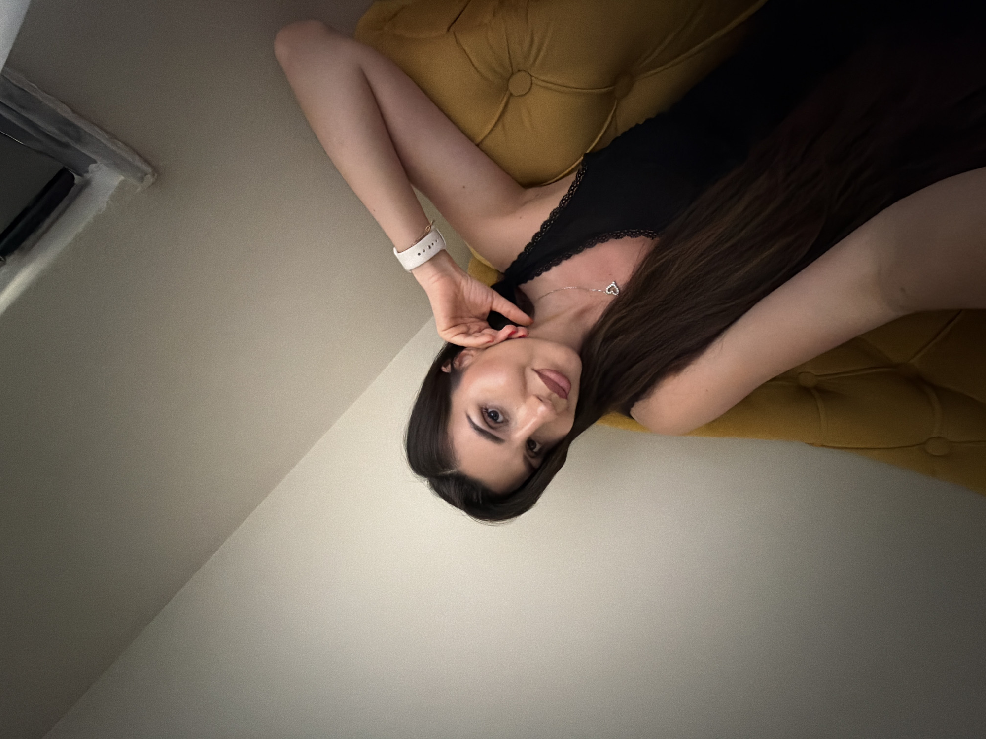 A young woman with long, dark brown hair is depicted lying on her side or back on a mustard yellow, tufted piece of furniture, likely a sofa or armchair. She wears a black sleeveless top with lace trim along the neckline and a silver necklace featuring a heart-shaped pendant. On her left wrist, she has a white smartwatch, and her fingernails are painted in a dark red or burgundy shade.
She looks directly at the camera with a calm, subtle smile, her lips slightly parted. Her right hand is gently positioned near her face, with fingers touching her chin and cheek. The background consists of plain, light-colored walls, possibly cream or light grey. In the upper left, a partial view of a window frame or vent structure is visible. A soft diagonal shadow line is present on one of the walls, indicating an indoor setting with ambient lighting. The scene is set indoors in Budva, Montenegro. There is no visible text in the image.