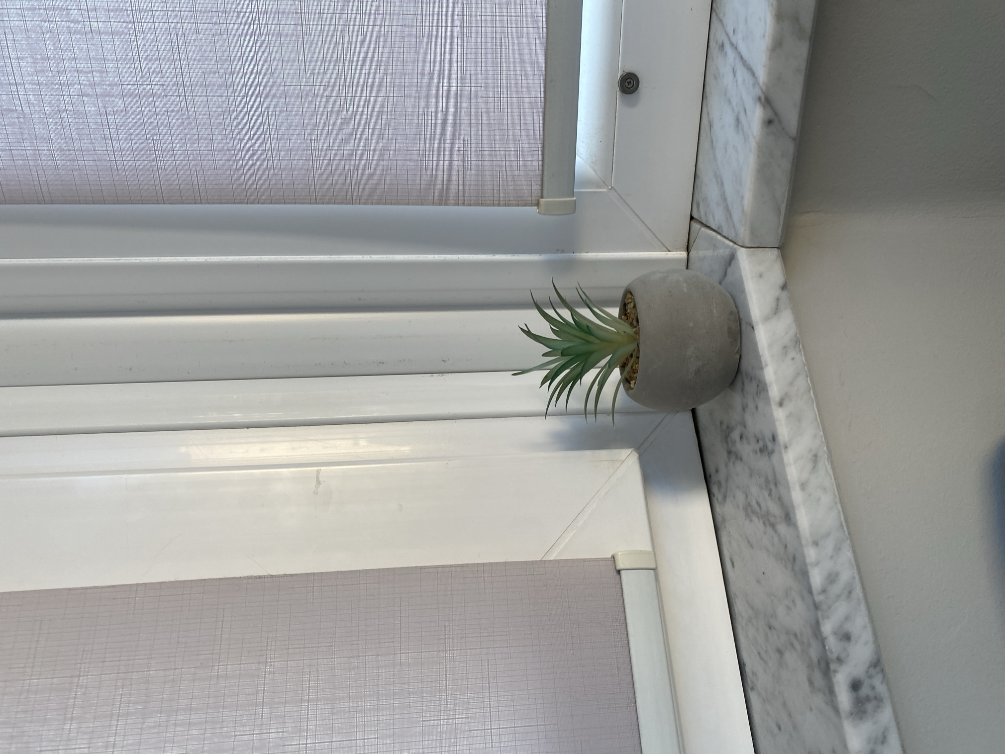 A small, artificial succulent plant in a grey, round pot sits on a marble windowsill. The windowsill is positioned in the corner of a window, with white window frames and textured, light purple roller blinds visible. The blinds are partially lowered, obscuring the view outside. The scene is well-lit, suggesting it is daytime. No people or animals are present. The image appears to be a close-up shot of the windowsill and plant. The location in Chișinău, Moldova, is not discernible from the visual information in the image.