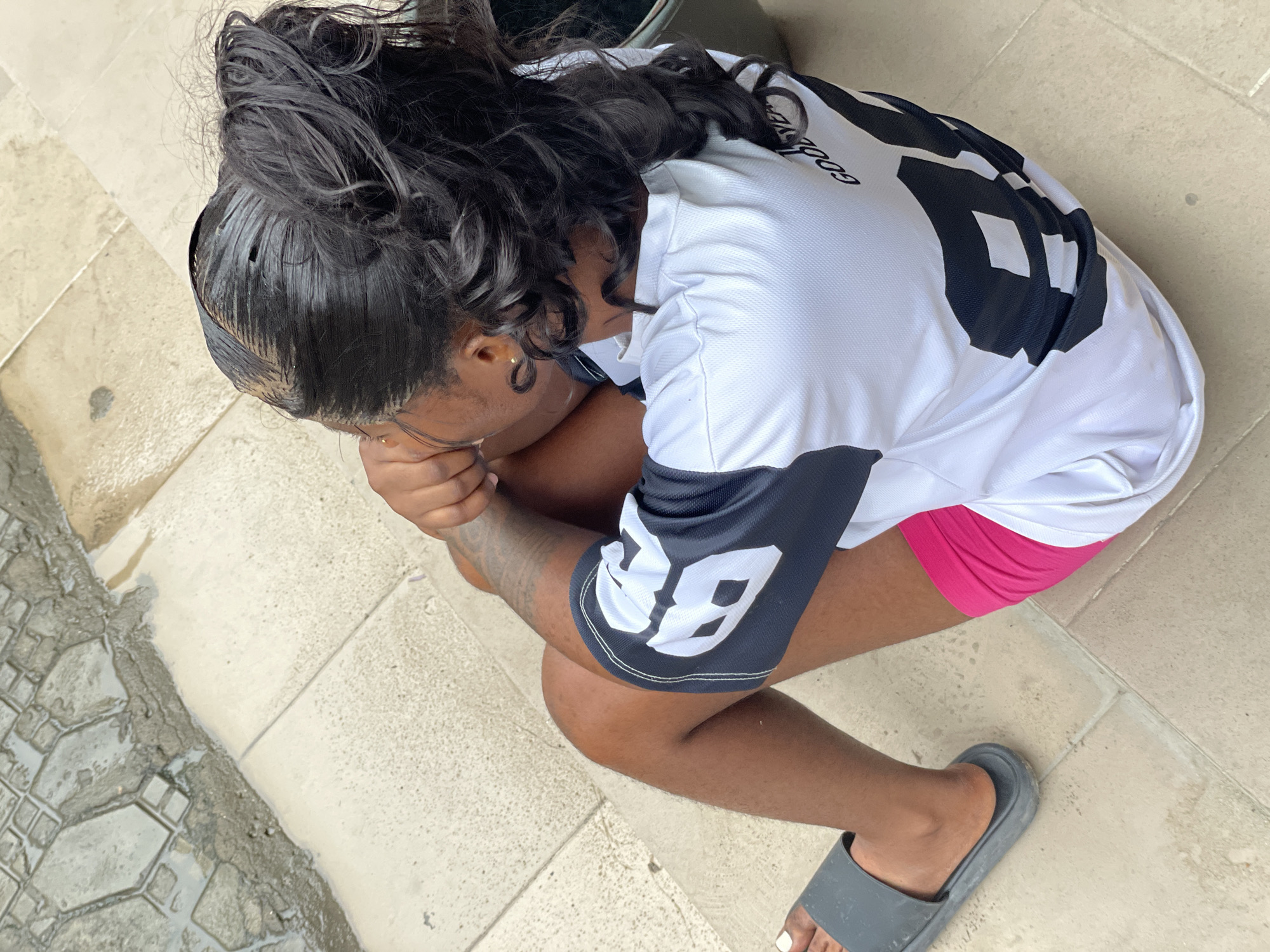 A top-down, medium shot depicts a dark-skinned person with black, curly hair, seated on a tiled surface. The person is wearing a white and black athletic jersey with the number "38" on the sleeve and pink shorts. They are also wearing a grey sandal on one foot. The person is crouched with their head lowered and arms wrapped around their legs. The background shows a tiled floor or walkway and a glimpse of wet pavement. The scene appears to be located outdoors. There are no other visible individuals or landmarks to determine the specific location within the city of Ado, Nigeria.