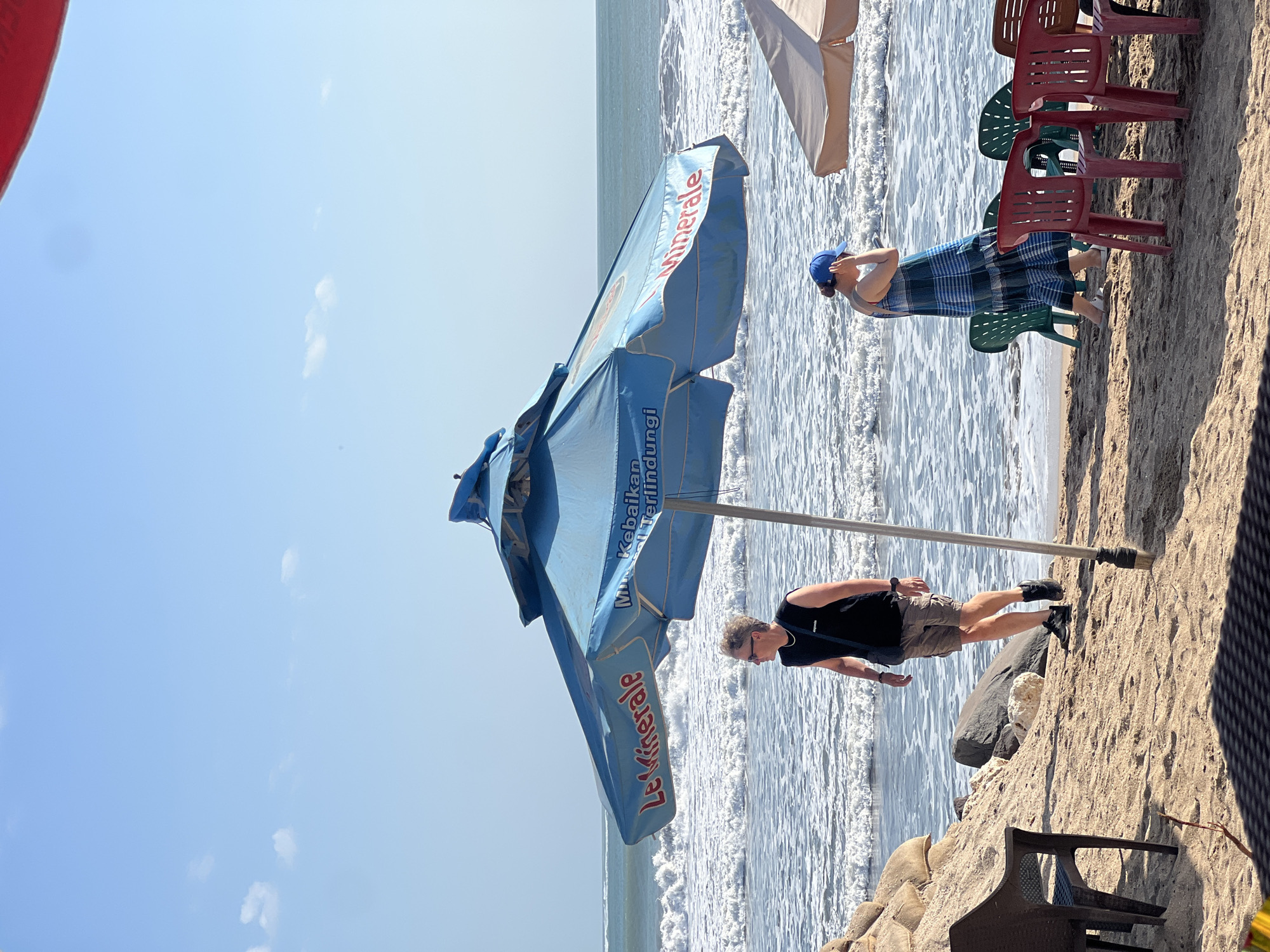 The image depicts a sunny day at Kuta Beach, Indonesia. In the foreground, a blue beach umbrella with "Le Minerale" branding is open, casting a shadow over the sand. A man wearing a black sleeveless shirt, khaki shorts, and black sneakers is standing near the umbrella, looking out towards the ocean. Behind him, a woman in a blue and white checkered dress and a blue baseball cap is walking on the beach, holding her hand to her head. Several plastic chairs in red and green are arranged on the sand, some of which appear to be associated with beachside food or drink vendors. The ocean in the background has moderate waves, and the sky is clear with a few scattered clouds. The overall scene conveys a relaxed atmosphere typical of a beach resort.