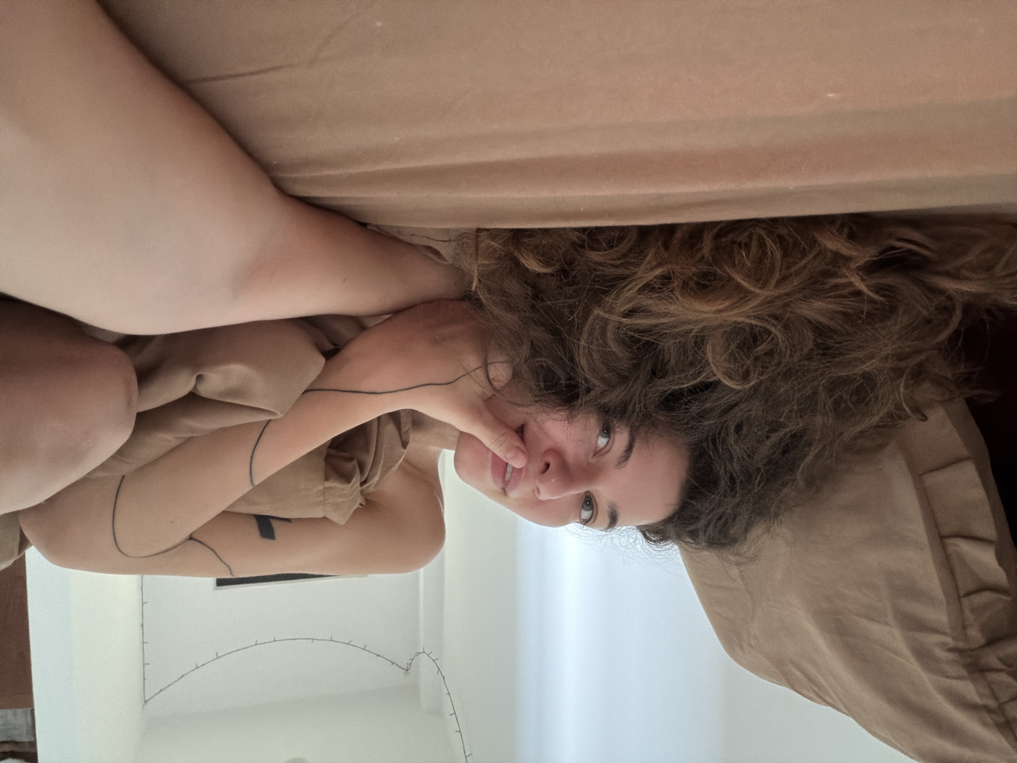 A woman with curly brown hair is lying in bed, looking at the camera. She is wearing a beige shirt and has line tattoos on her arm. The bed is covered with a beige sheet. A string of lights is visible in the background, which appears to be a white wall. The location is an interior space, presumably a bedroom. There is no indication this image was taken in Đà Nẵng, Vietnam.