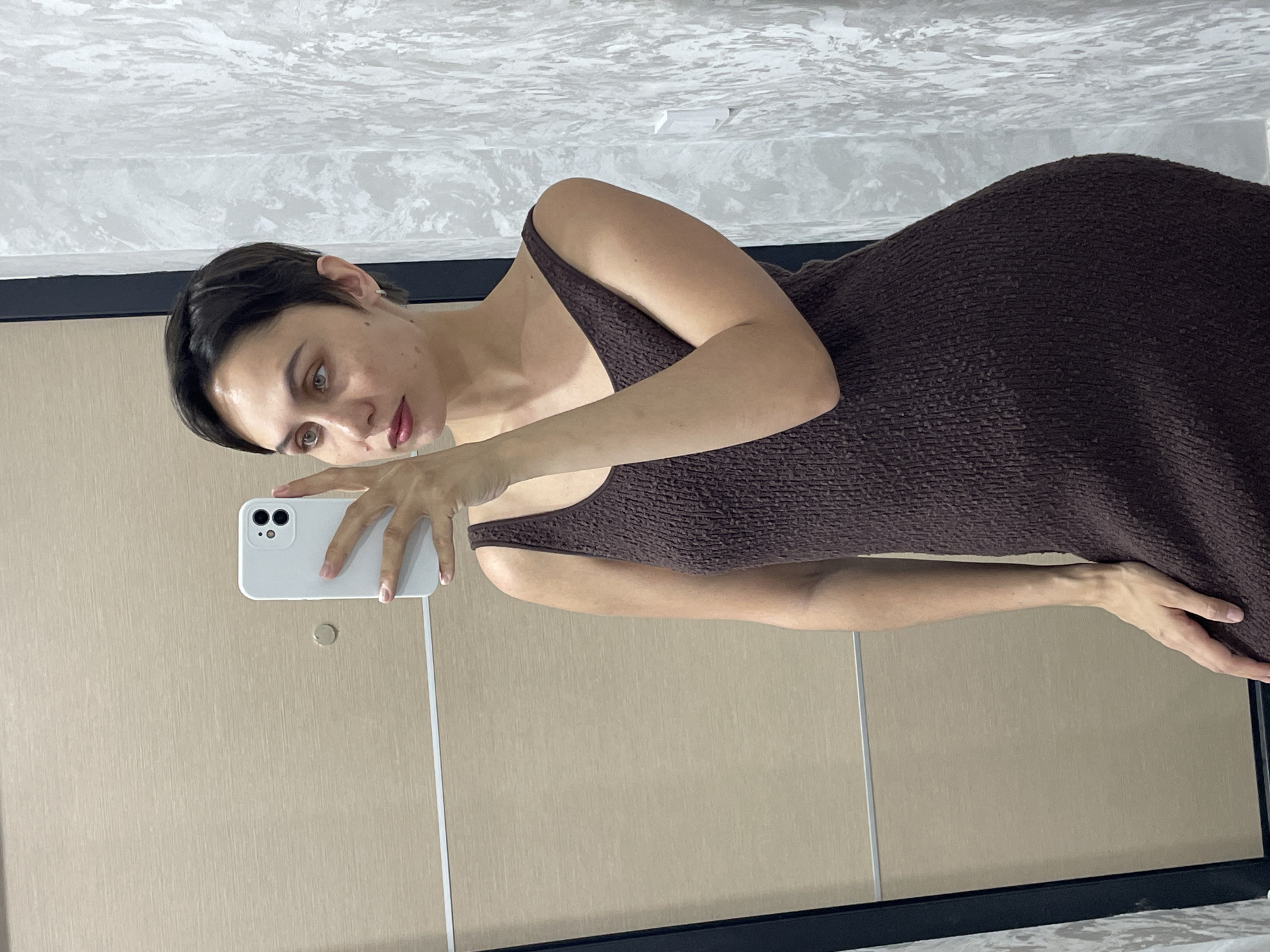 A woman is taking a selfie in front of a mirror. She is wearing a brown, sleeveless dress. The background includes a wall with a patterned surface and what appears to be a closet door. There is no information to place the location in Izhevsk, Russia.