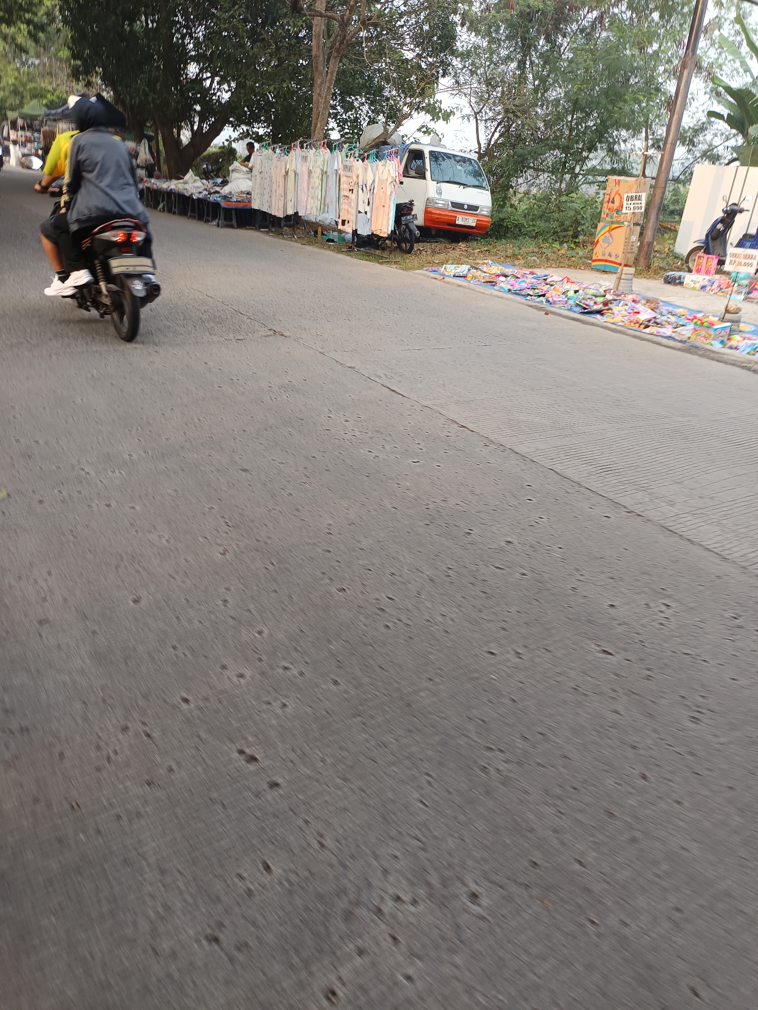 A motorcyclist, wearing a black helmet and a grey jacket over a yellow shirt, rides down an asphalt road in Sodong, Indonesia. The motorcyclist is seated on a dark-colored motorcycle, with the rear of the vehicle and a portion of the road visible. In the background, a row of clothing items is displayed on hangers, suggesting a market or roadside stall. A white mini-truck is parked nearby, and further down the road, various colorful items are spread out on blue tarps, likely merchandise for sale. Green trees and foliage line the side of the road, and a utility pole stands tall. The scene appears to be an outdoor commercial area, possibly a street market.