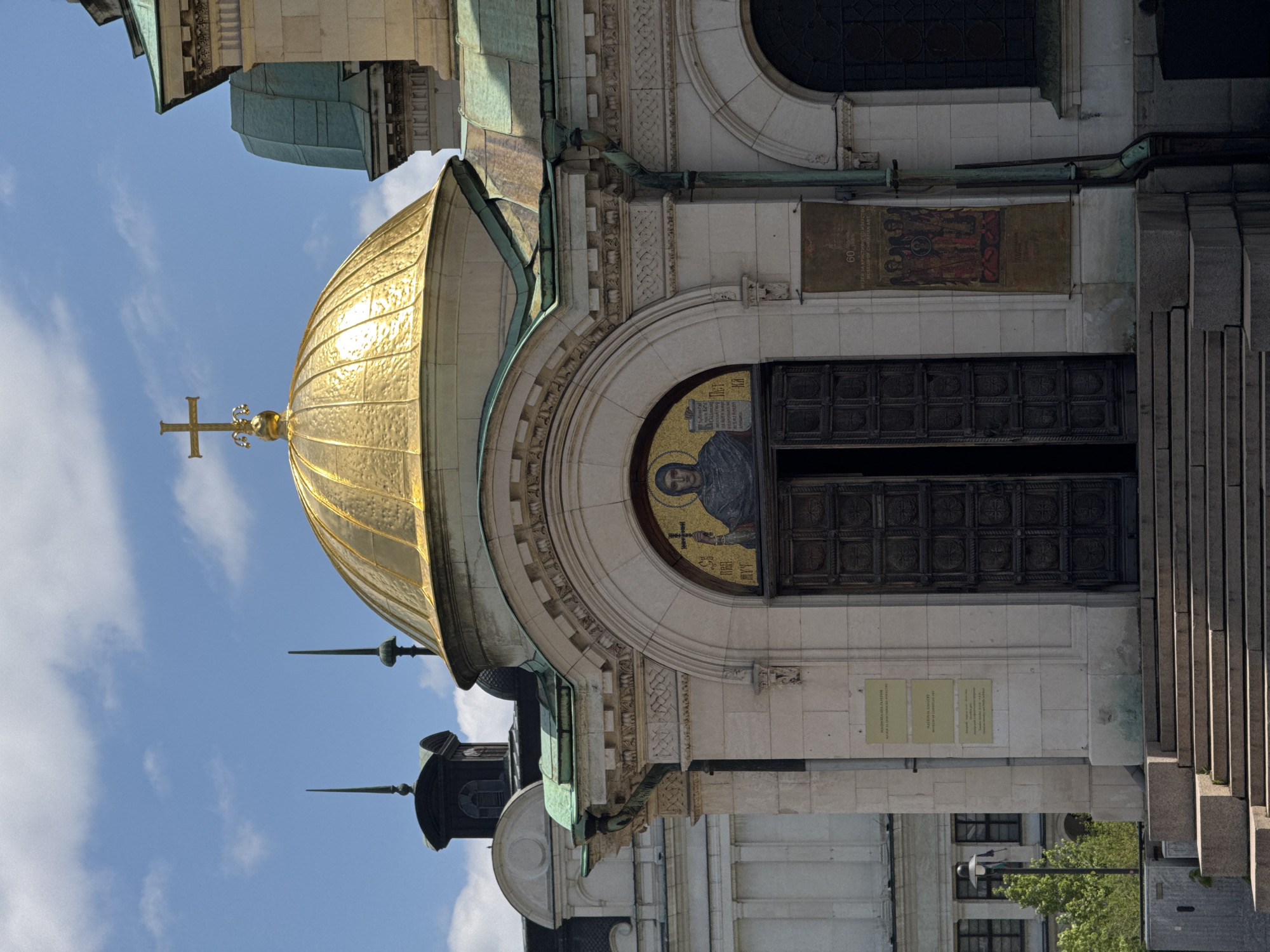 The image depicts the Alexander Nevsky Cathedral in Sofia, Bulgaria. Visible is a golden dome topped with a cross against a blue sky. A mosaic of a saint with a cross adorns the arched doorway. A partially open door stands to the right of the mosaic. Plaques are visible on the wall near the door. A green roof is visible below the golden dome. Steps lead up to the building entrance on the right.