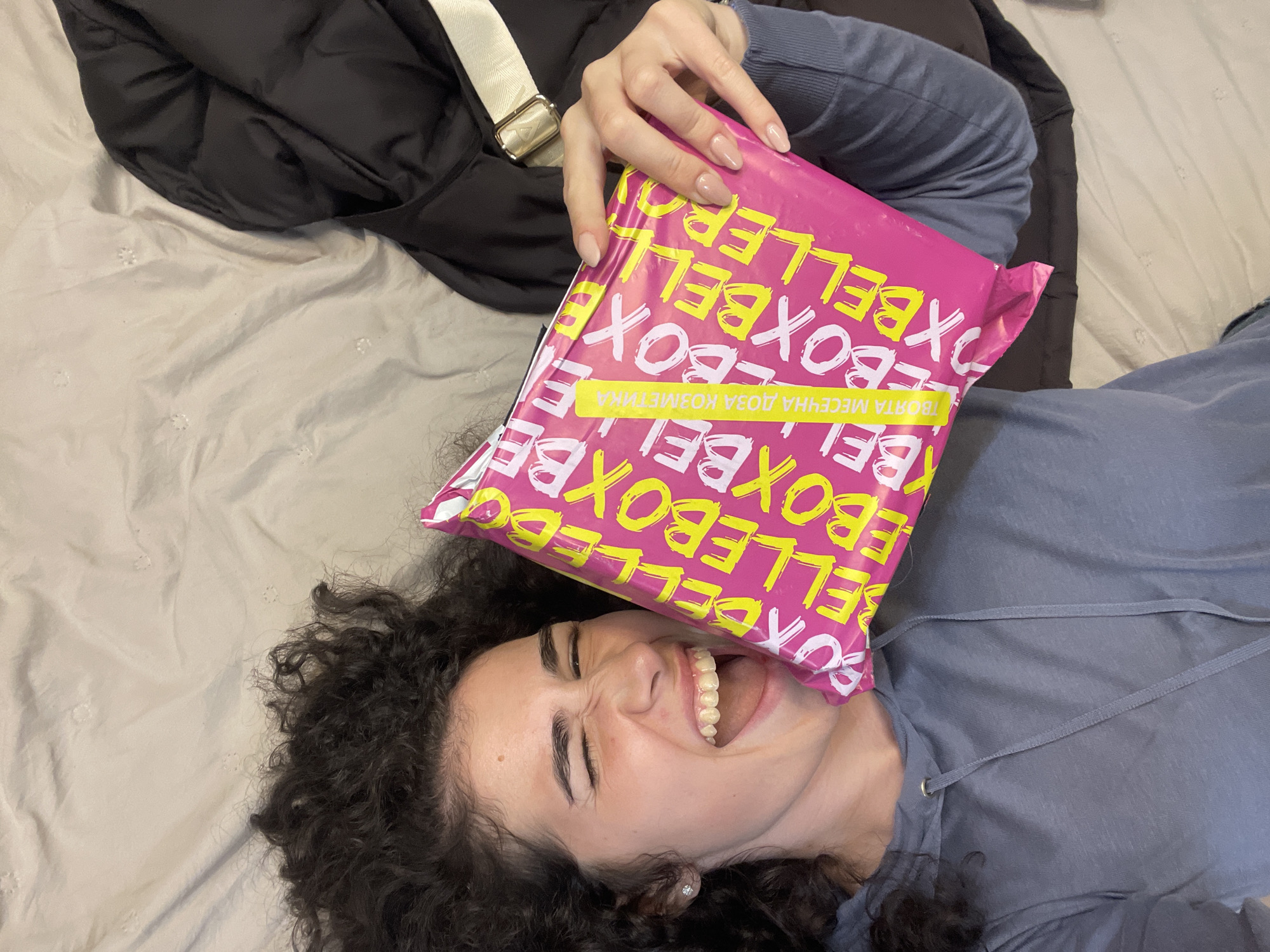 A young woman with dark, curly hair lies on her back on a light grey bedspread, laughing joyfully. She is holding a vibrant pink package with her right hand, placing it partially over her face. Her eyes are squeezed shut in amusement, and her mouth is wide open, revealing her teeth and what appears to be a clear aligner or retainer. She wears a blue-grey long-sleeved top with a drawstring neckline and has light-colored nail polish.
The pink package is covered with repeating text in yellow and white, reading "BELLEBOX" and "BOXBELLER." A prominent yellow strip across the package displays white text in Bulgarian: "ТВОЯТА МЕСЕЧНА ДОЗА КОЗМЕТИКА," which translates to "Your Monthly Dose of Cosmetics." This indicates the package contains cosmetic products.
Beside her, a black puffer jacket or coat and a light-colored shoulder bag with a gold buckle are visible. The setting appears to be indoors, likely a bedroom in Sofia, Bulgaria, given the provided context. The activity captures a moment of happiness and excitement as she seemingly interacts with her new delivery.