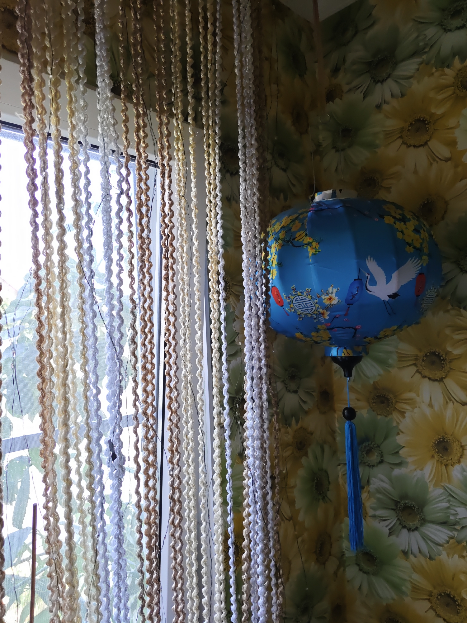 The image shows a corner of a room with floral wallpaper. A blue paper lantern decorated with cranes, flowers, and other oriental motifs hangs from the ceiling. A beaded curtain, made of strands of various colors and textures, hangs in front of a window on the left side of the image. The wallpaper features large, stylized flowers in shades of yellow and green, resembling sunflowers or chrysanthemums. The lantern has a blue tassel hanging from its bottom. There is no visible information in the image that places the scene in Rostov-on-Don, Russia.