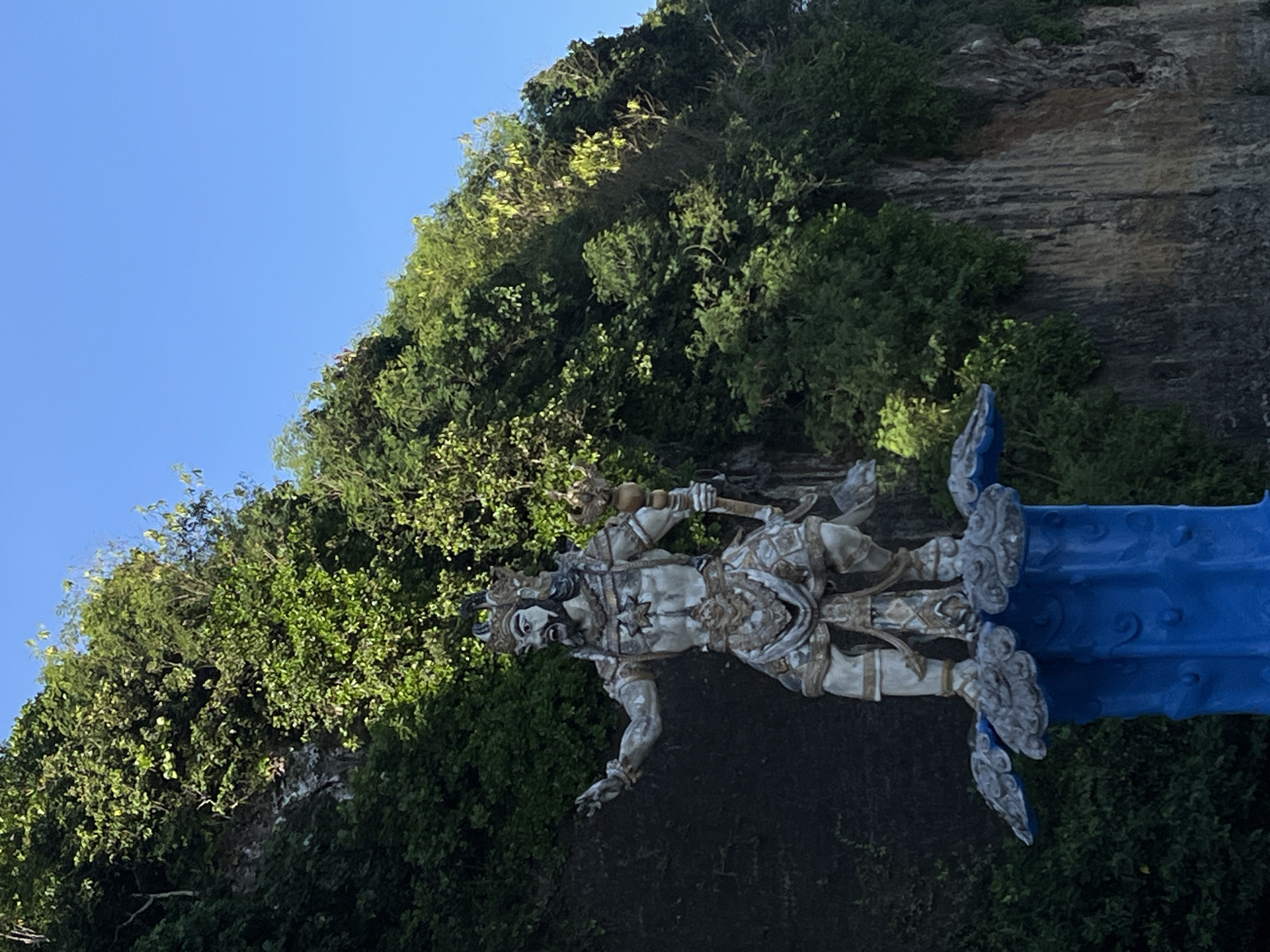 The image depicts a statue of a deity, possibly Hindu, positioned against a backdrop of lush green vegetation and a rocky cliff. The statue is predominantly white or light-colored, with intricate carvings and details on its clothing and accessories. It appears to be holding a scepter or weapon. Part of a blue structure is visible behind the statue. The location is Kuta Selatan, Indonesia.