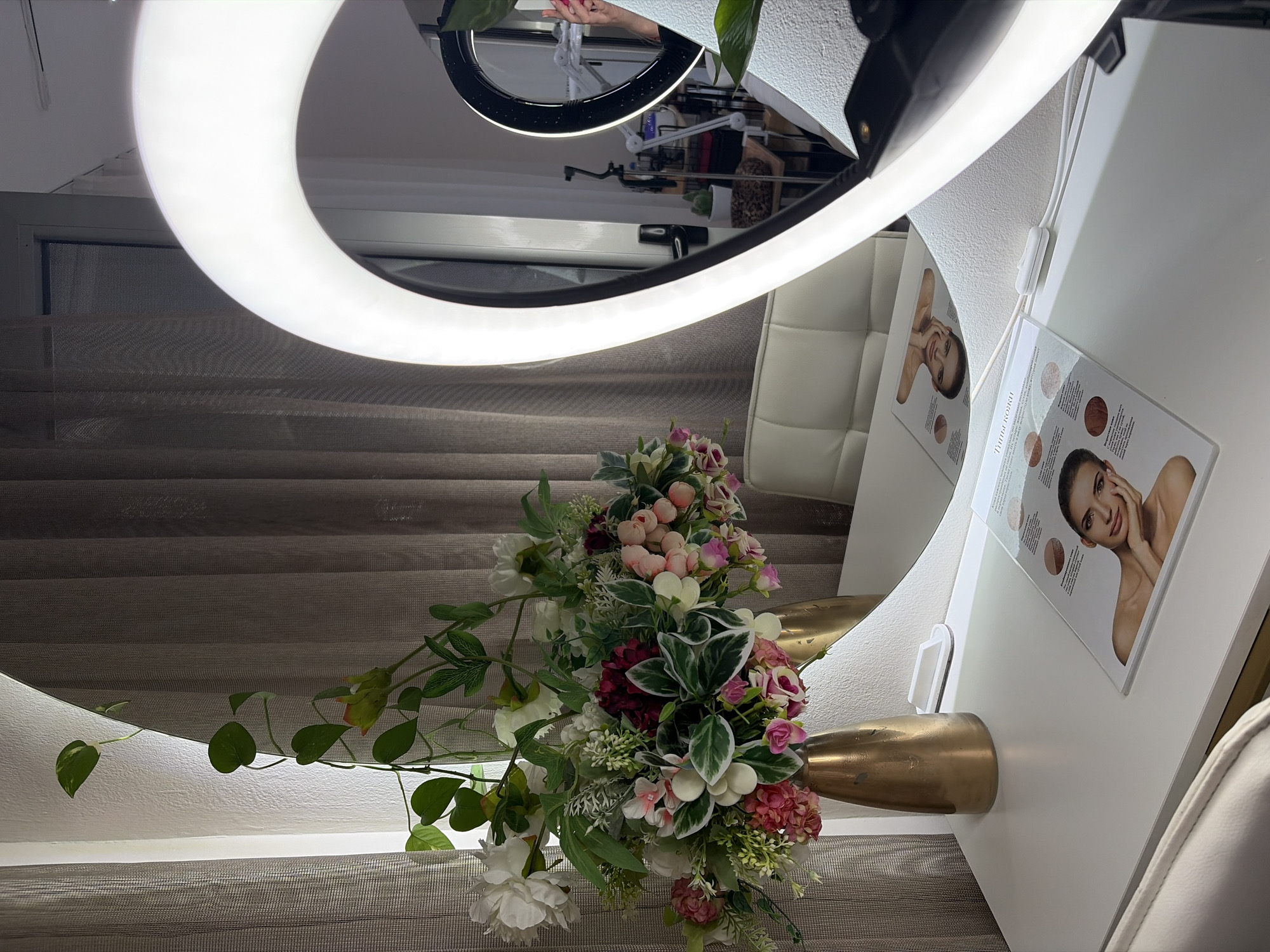 The image depicts an indoor scene, likely a vanity or makeup station, with a large, illuminated ring light prominently featured. A white desk or table surface occupies the foreground, holding a vase of artificial flowers, a small white switch, and a glossy brochure displaying images of women's faces and what appear to be skin tone charts.
To the left, a trailing green plant with heart-shaped leaves adds a touch of organic decor. The background consists of translucent beige curtains and a wall. The large mirror reflects other elements of the room, including another ring light, various studio equipment such as a tripod and lighting stands, and a white upholstered chair. A person's hand holding a phone is visible in the reflection of the second ring light. The overall impression is one of a professional setup for beauty or content creation. The image does not contain any explicit visual cues that definitively place it in Budva, Montenegro.