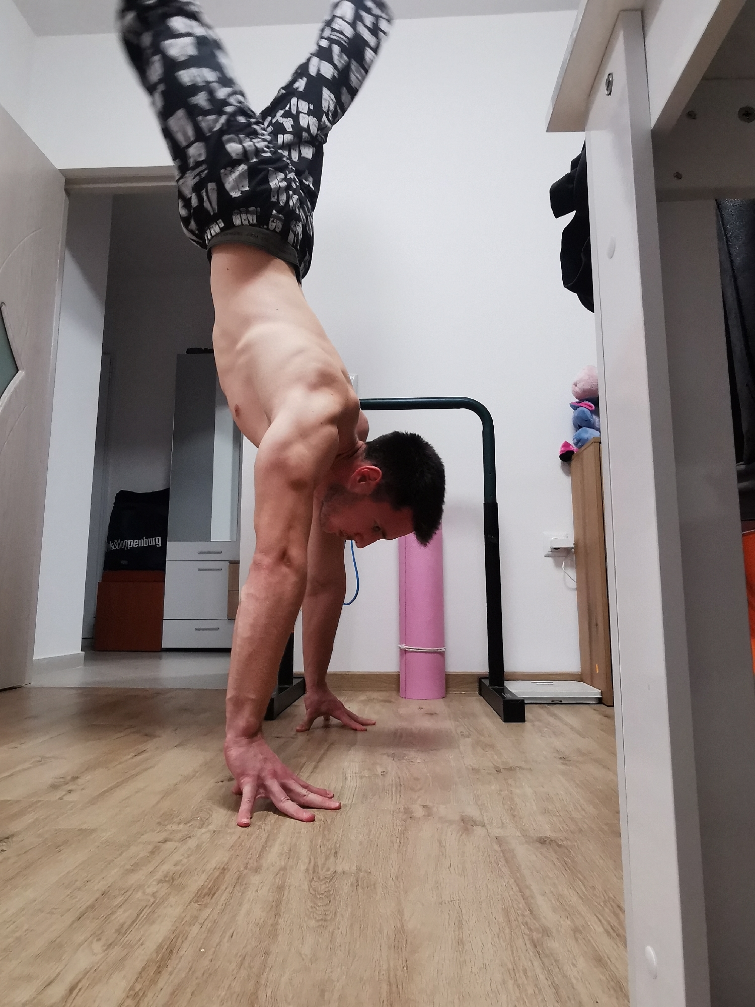 A man is performing a handstand in an indoor setting. He is wearing patterned pants and is shirtless. His legs are split in a V-shape above his body. He is facing a pull-up bar set up next to a rolled up pink yoga mat. A white scale is placed on the floor near the yoga mat. A white cabinet and a black bag are visible in the background. The floor is wooden. The location is likely an apartment or home gym in Burgas, Bulgaria, given the local context.