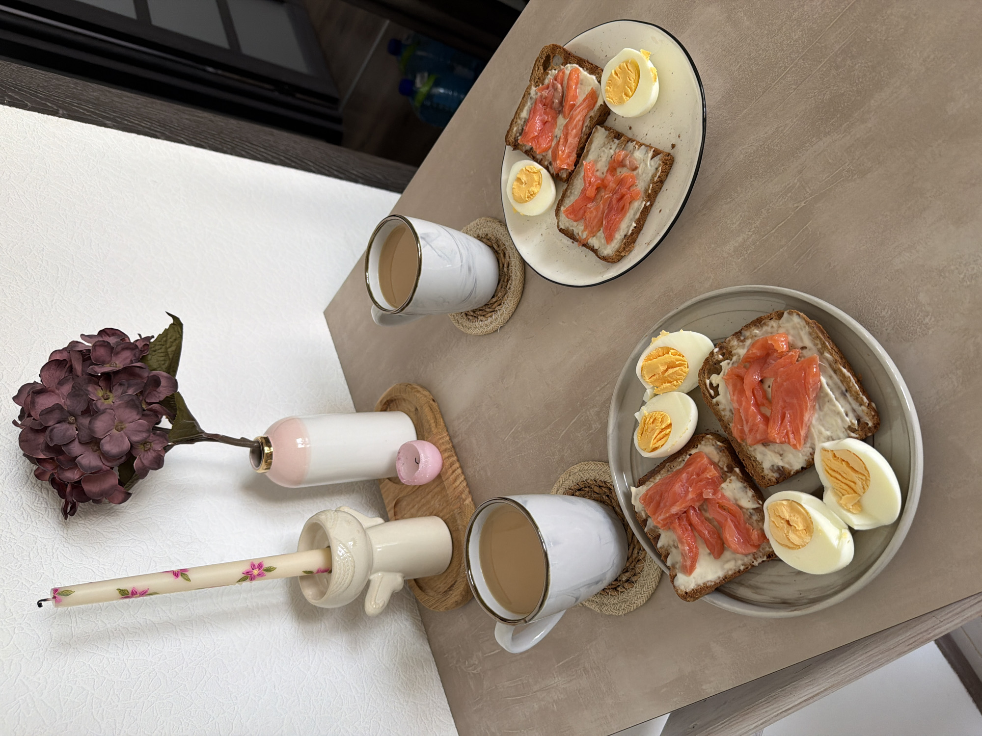 The image shows a breakfast table set for two. On the table, there are two plates of open-faced sandwiches with salmon and cream cheese, and sliced hard-boiled eggs. There are also two mugs of coffee with milk. In the background, there is a decorative candle in an angel-shaped holder and a vase with a dried flower arrangement. The table appears to be in a residential setting in Kyiv, Ukraine.