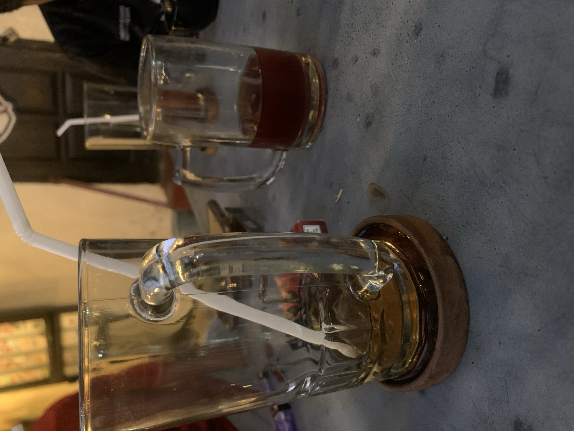 The image captures a close-up, eye-level view of two glass mugs on a textured, gray surface, likely a table. Both mugs contain a light brown liquid and have white plastic straws. The mug in the foreground is larger and appears to be a beer mug, with a thick handle. It is mostly empty, with liquid only in the bottom. A wooden coaster sits beneath it. The second mug, to the right and slightly behind, is smaller and shows more liquid. The background is blurred, suggesting an indoor setting with warm lighting. Elements in the background include what appears to be a dark wooden door or wall, and a shelf with multiple items on it, some of which are rectangular and appear to be boxes or books. The overall atmosphere suggests a casual gathering or dining experience. The location of Kraton, Indonesia, is not directly depicted in the image but is provided as context for the scene.
