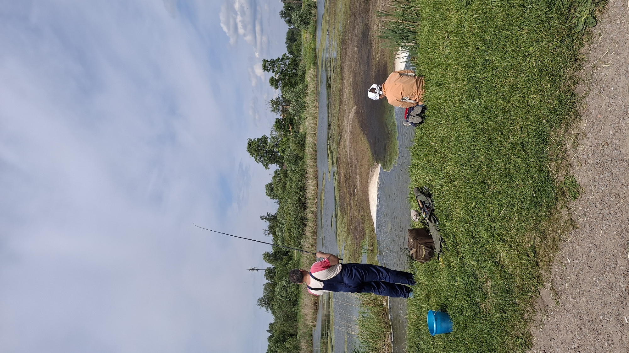 This is an outdoor scene captured during daylight in Krasnoye Selo, Russia, depicting two individuals engaged in fishing by a body of water.
The upper portion of the image shows a partly cloudy sky with patches of light blue interspersed with scattered white stratocumulus clouds. Below the sky, a dense line of green deciduous trees and shrubs forms the horizon, suggesting a natural or semi-natural environment. A cellular transmission tower is partially visible above the trees on the far left.
The central feature is a wide, shallow body of water, possibly a pond or slow-moving river, which appears largely covered with green aquatic vegetation, such as algae or duckweed, giving it a somewhat murky appearance. There are clearer patches of water that reveal a brownish sediment or bottom. A distinctive lighter-colored, crescent-shaped sandy or muddy area extends into the water from the near bank, approximately in the center of the frame.
In the foreground, a vibrant strip of mown green grass separates the water's edge from a gravel path or dirt road that runs across the bottom of the image.
Two male subjects are positioned on the grassy bank, facing the water.
On the left, a man of medium build stands with his back to the camera. He wears dark blue overalls over a light-colored (possibly white or light grey) t-shirt with red trim around the collar and sleeves, and blue athletic shoes. He holds a long, dark fishing rod with both hands, extending it over the water, appearing to be actively fishing or casting.
To his right, a second man is seated on the grass, also with his back to the camera. He wears an orange long-sleeved shirt, dark trousers, and a white bandana or cloth covering his head. His shoes are visible. He appears to be observing the water or waiting.
Between the two subjects, various fishing equipment is laid out on the grass: a bright blue plastic bucket on the far left, a brown canvas or fabric tackle bag (partially open) in the center, and a long, dark zippered case (likely a rod case) next to it. Some small, unidentifiable items are scattered near the tackle bag.