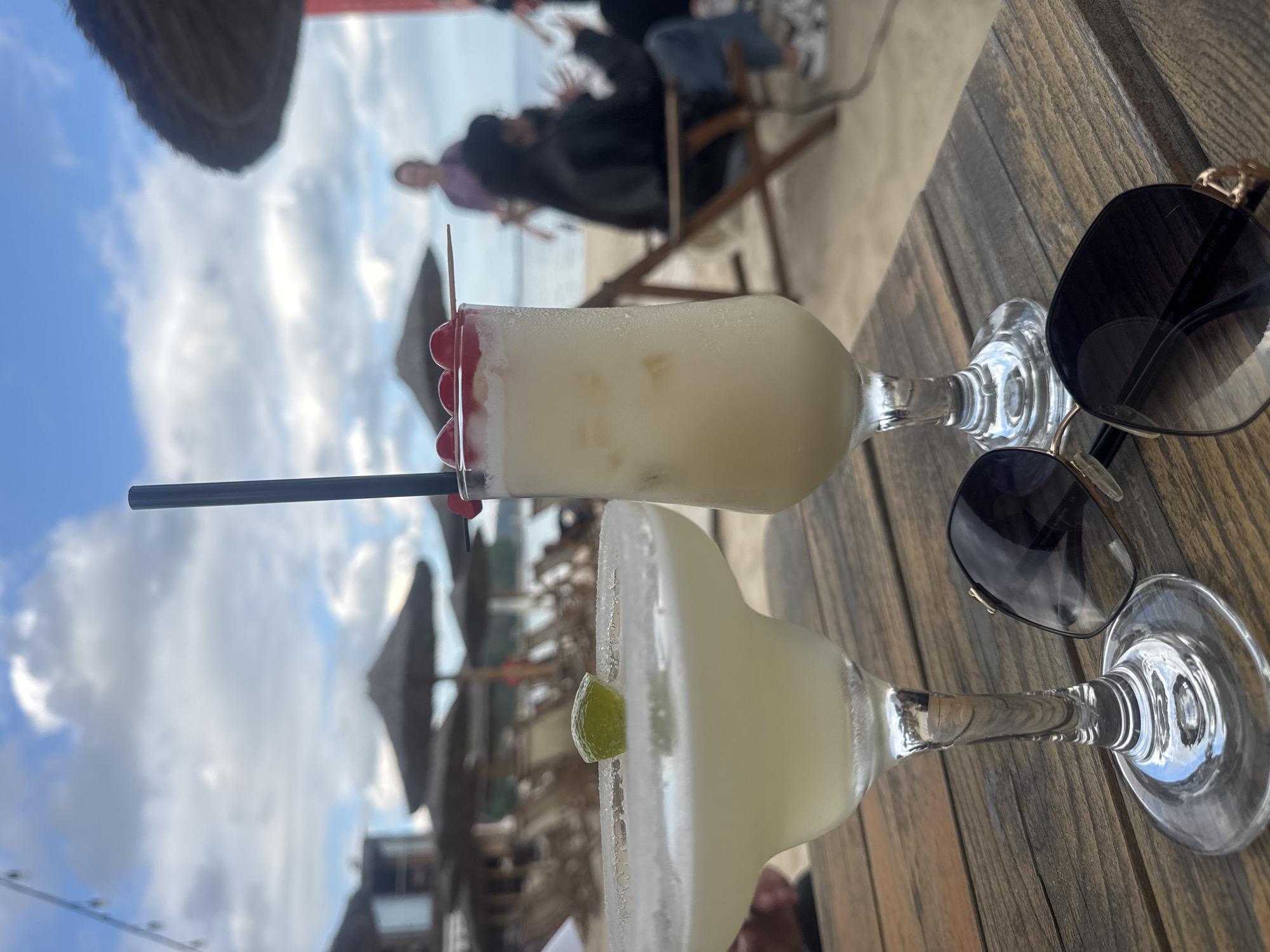 The image presents two cocktails on a wooden table, likely at a beachside location. The cocktail closest to the viewer appears to be a margarita, with a salted rim and lime garnish. The other cocktail, positioned behind it, is a white beverage garnished with cherries and a black straw. A pair of sunglasses rests on the table beside the drinks.
In the background, a beach scene unfolds with people relaxing under straw umbrellas. Based on the location indicated, it is likely that this scene is in Varna, Bulgaria, situated along the Black Sea coast. The sky is visible above, with patches of blue and clouds.