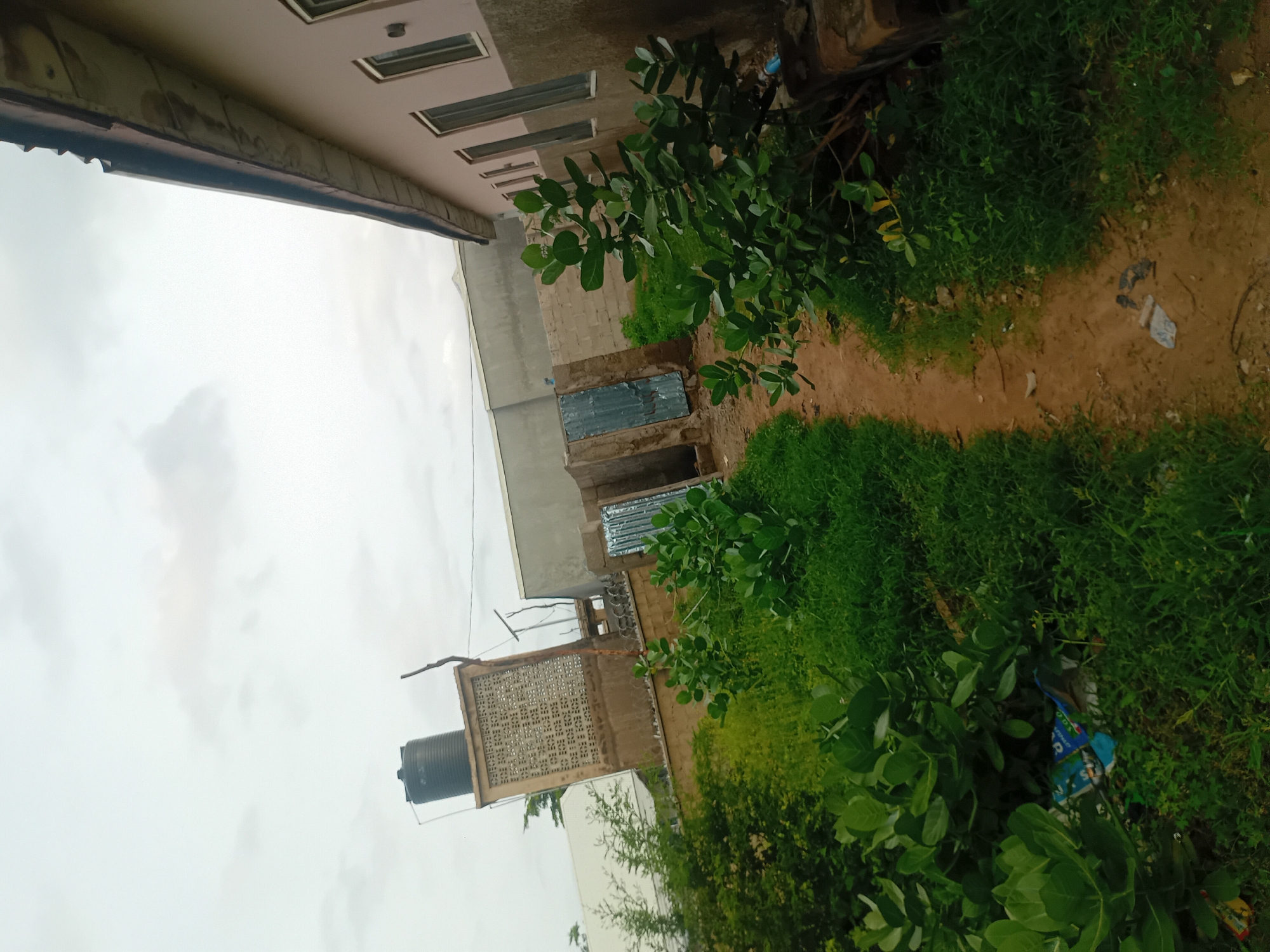 The image depicts an outdoor scene, likely a residential area in Jauro Yinu, Nigeria. A narrow, unpaved dirt path runs diagonally through the frame, bordered by lush green vegetation and grass. To the right, a light-colored building with a dark roof and multiple windows is visible, suggesting a dwelling or commercial structure. On the left side, a portion of a brown fence is seen, with more dense greenery behind it. Further in the background, a tall, rectangular structure with a patterned facade and a black water tank on top is present, indicating typical infrastructure in the area. There are also glimpses of other buildings and structures, including a corrugated metal gate. The sky is overcast with clouds. The scene appears to be devoid of people, focusing on the built environment and natural elements.