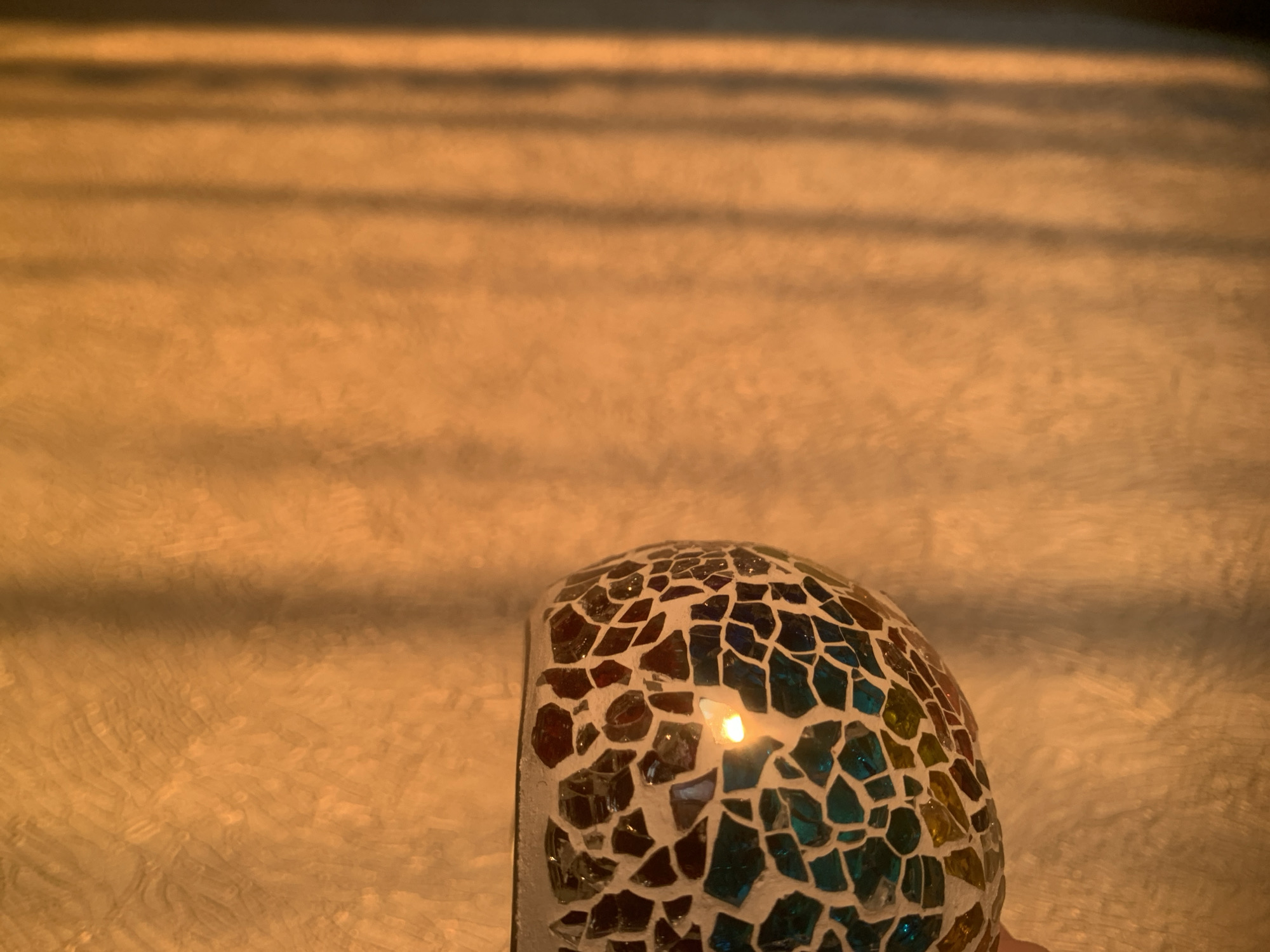 In a location identified as Izhevsk, Russia, an indoor scene is depicted under warm, orange-yellow illumination. The primary subject is a hemispherical or spherical object, approximately 15-20 cm in diameter, positioned in the lower-left portion of the frame. This object features a mosaic surface composed of irregularly shaped fragments in teal, amber, and burgundy colors, set within a light-colored matrix. A bright point of light is reflected or emitted from its surface. A human hand, specifically a thumb and index finger, is partially visible, gripping the object from its lower-left side. The background consists of a textured wall exhibiting a subtle, irregular pattern. Multiple distinct, parallel vertical shadows are cast across the right side of the wall, indicative of light filtering through a structured obstruction, such as window blinds or a grate. No other individuals or objects are present in the visible area.