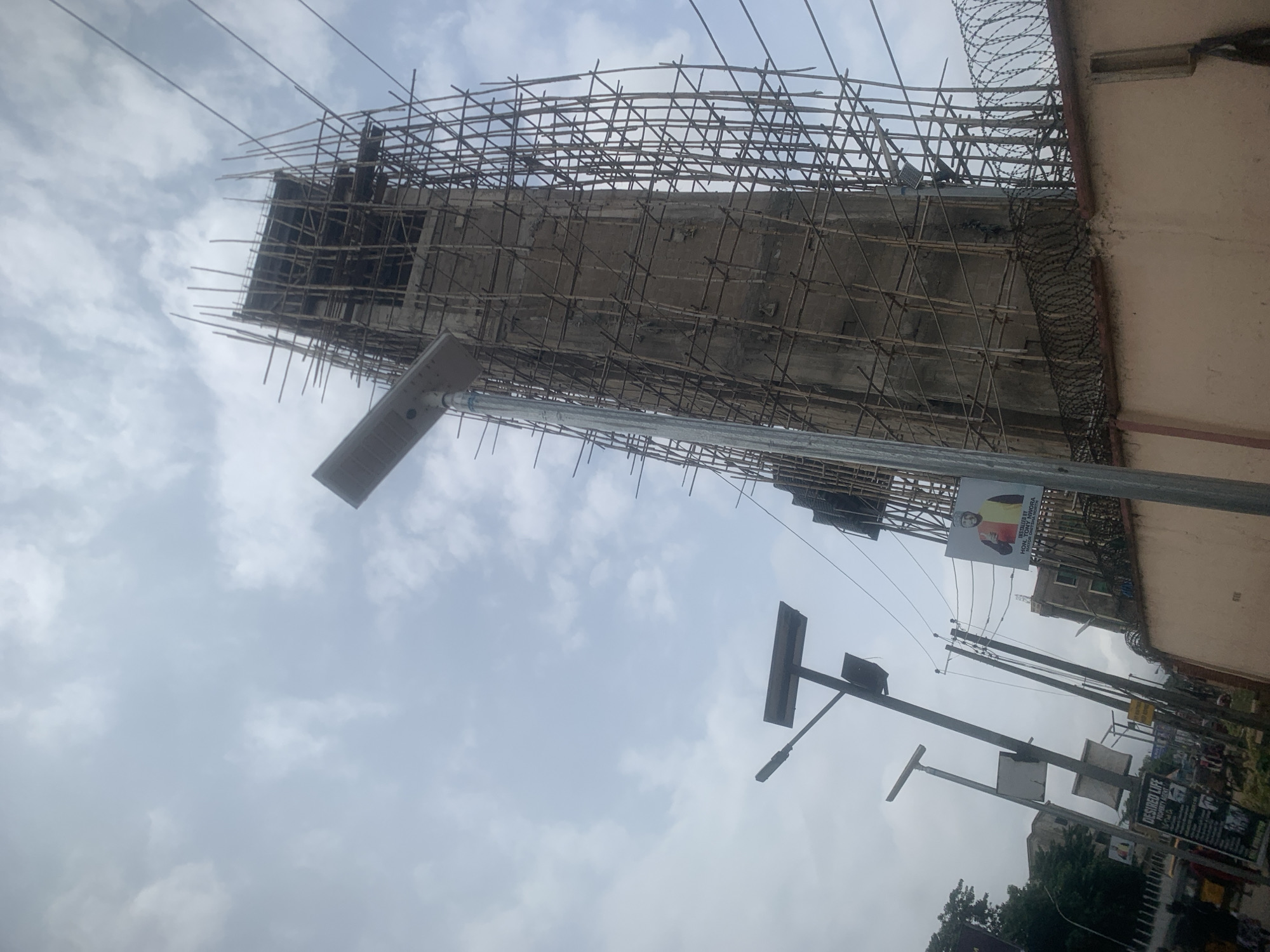 The image depicts a tall, unfinished concrete tower under construction, heavily encased in bamboo scaffolding, on a cloudy day in Onitsha, Nigeria. The tower appears to be a prominent structure within the urban landscape. In the foreground, a modern street lamp with a solar panel is visible, casting a shadow. Further to the left, other street lights and poles with solar panels are present. A section of a building with a light-colored wall, topped with barbed wire, runs along the bottom right of the frame. A large banner featuring a man in traditional attire, identified as "HON. DR. IYORY NORVA" and "GOVERNOR OF..." is affixed to the building's wall. The scene suggests an active construction site and a busy urban environment with street infrastructure and promotional materials.