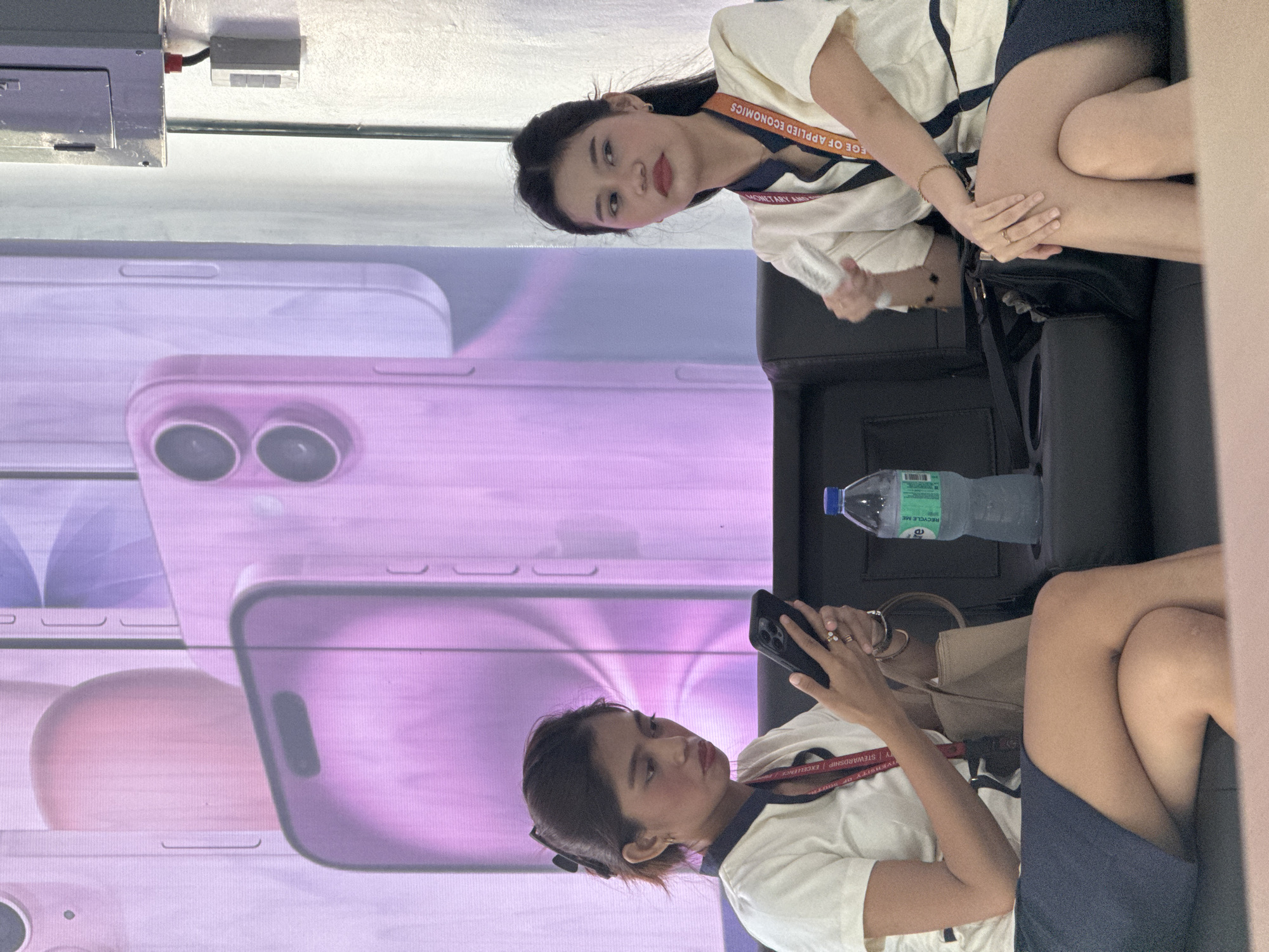 Two women are seated on a dark couch in front of a large, purple-toned display featuring images of smartphones. The woman on the left is looking down at her phone, which she holds in her right hand. She wears a white collared shirt with a dark trim and a dark skirt. A lanyard with text is visible around her neck, and she has a handbag on her lap. The woman on the right is looking to her right, away from the camera, with her left hand near her face. She wears a similar uniform as the woman on the left, including a white collared shirt with dark trim and a dark skirt, and a lanyard. A bottle of water is placed in a cup holder between the two women. The environment appears to be an indoor exhibition or event space, likely in Davao City, Philippines, given the context provided.